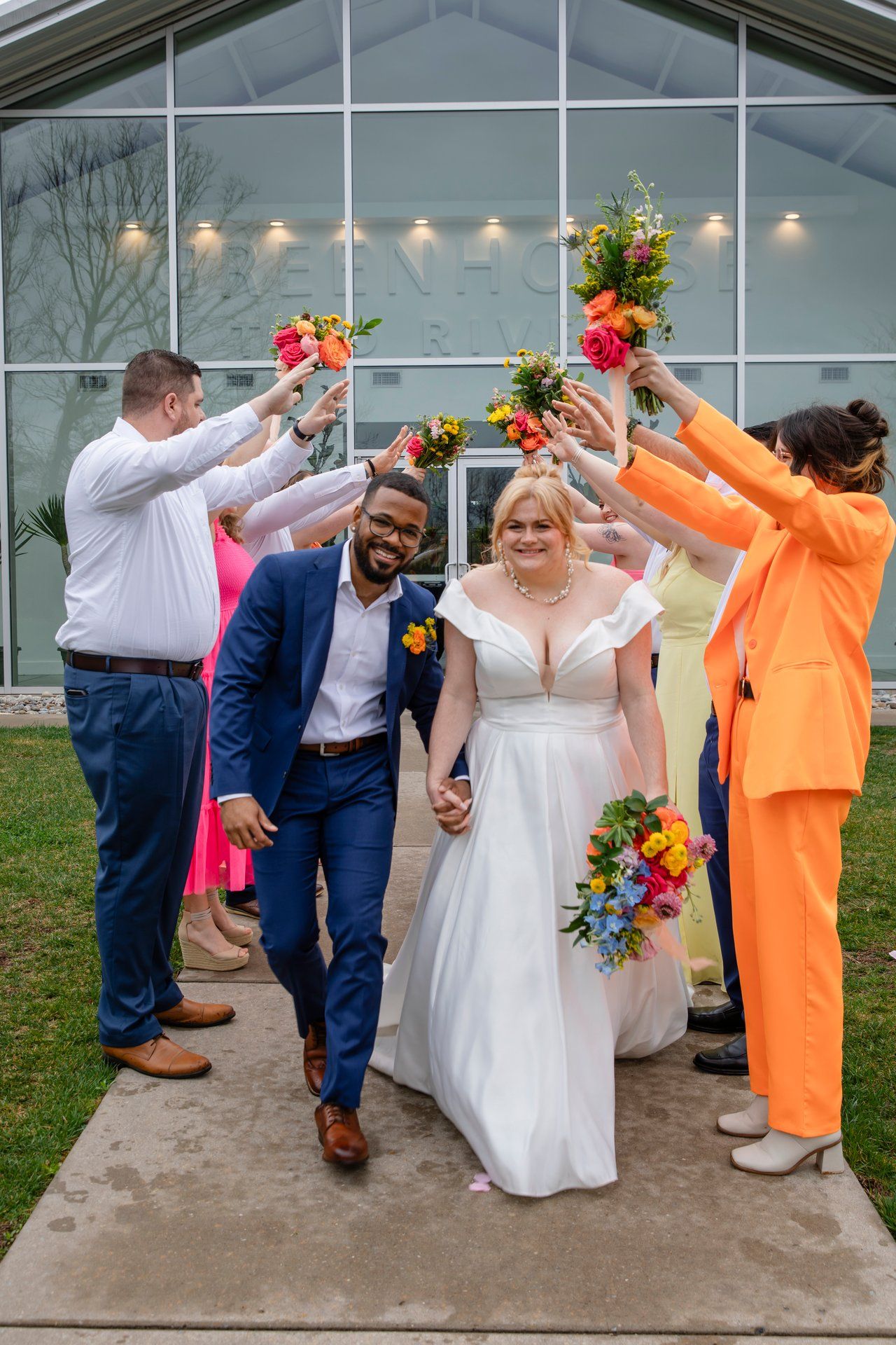 Wedding party celebrates by raising colorful bouquets as newlyweds walk down path.