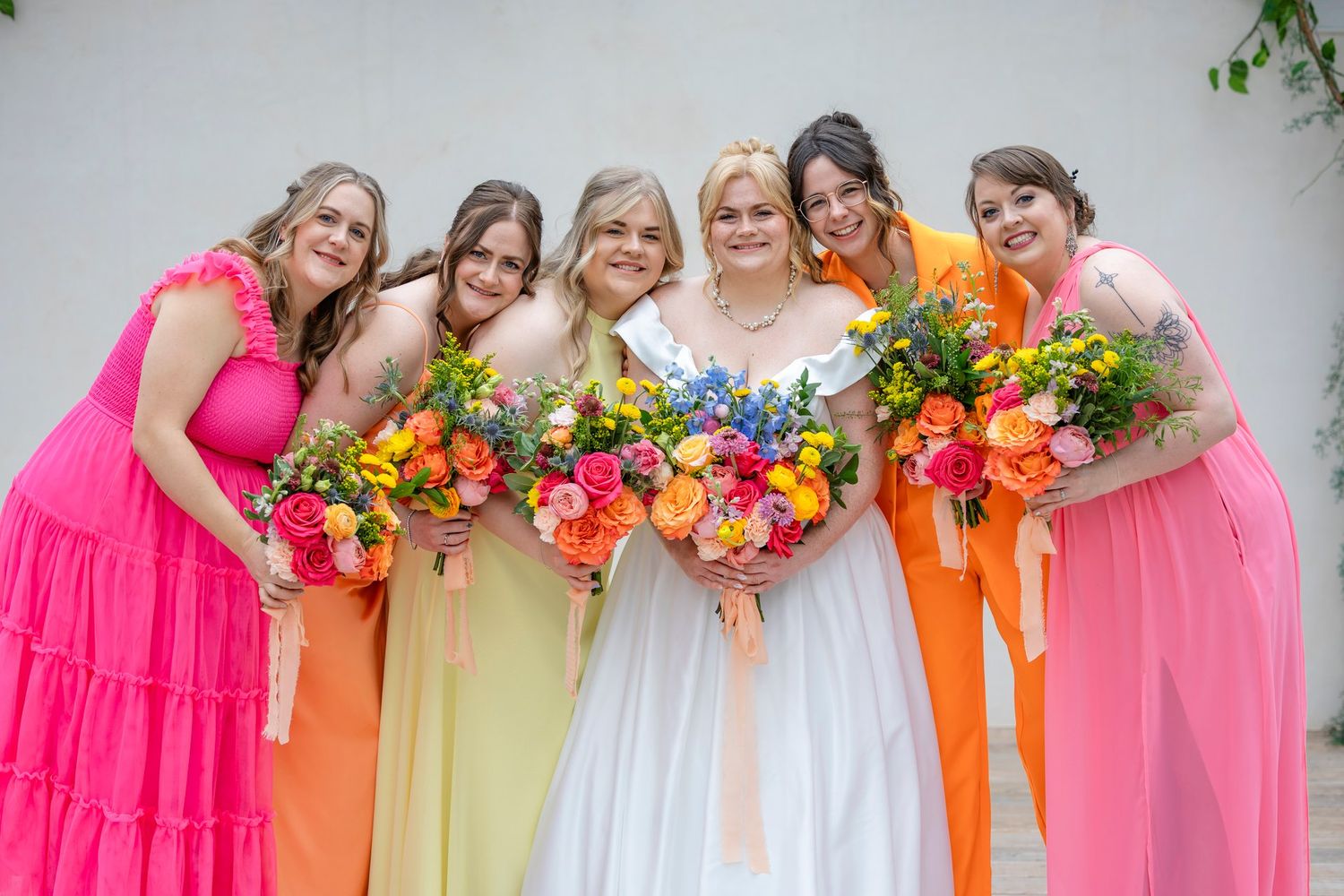 A colorful bridal party in bright pink and orange dresses poses together while holding vibrant floral bouquets.