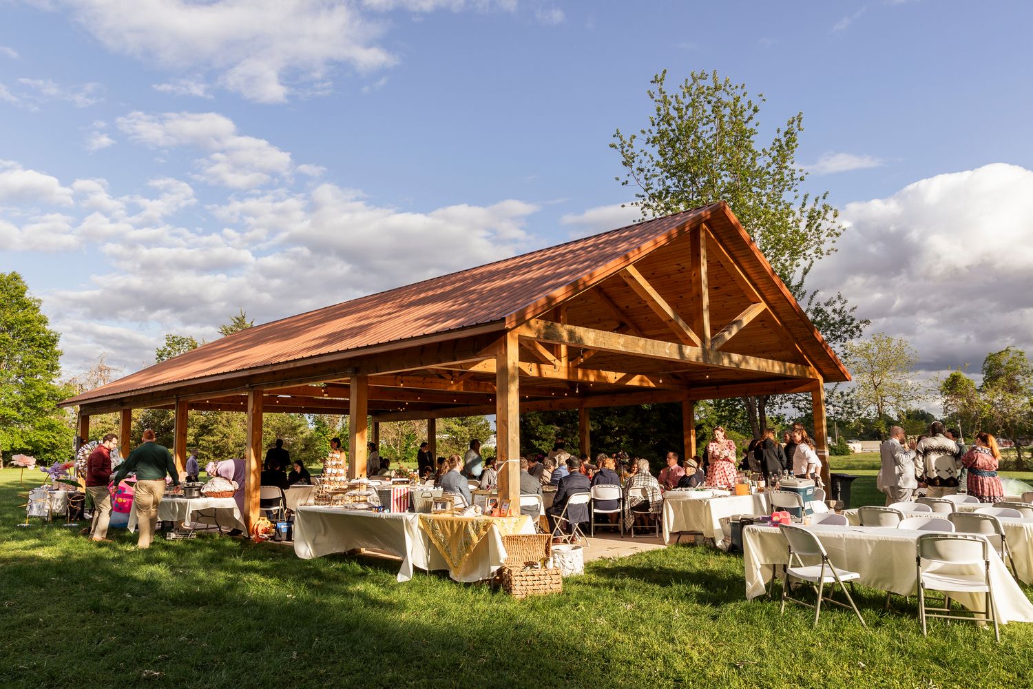 Outdoor wedding reception setup under wooden pavilion with white tables and chairs on a sunny day.