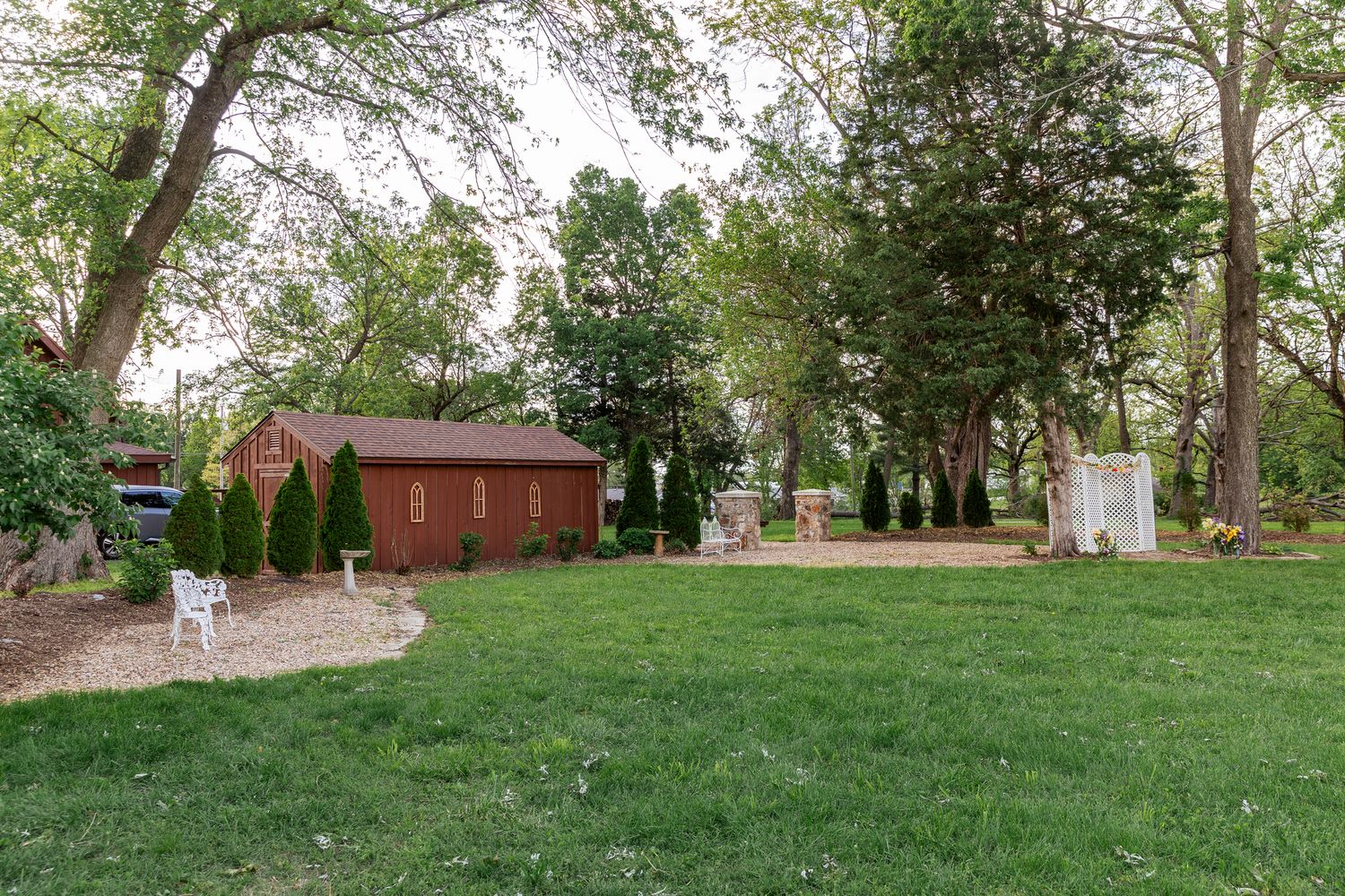 A well-landscaped backyard with a red wooden shed, tall trees, and manicured lawn surrounded by garden beds.
