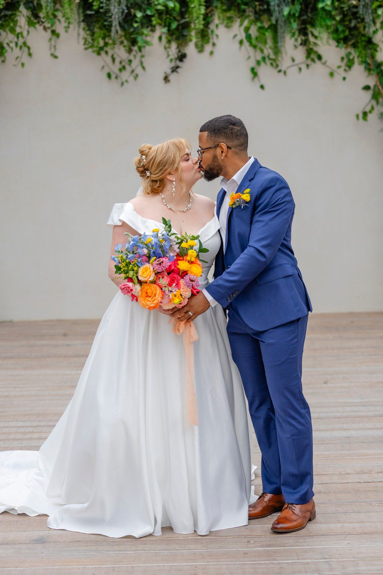 A wedding couple shares a romantic moment surrounded by hanging greenery with a vibrant bouquet.