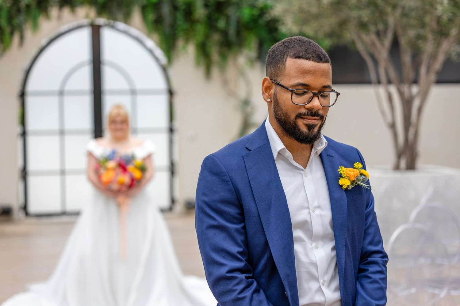 A groom in navy blue suit and glasses stands looking down during an outdoor wedding ceremony.