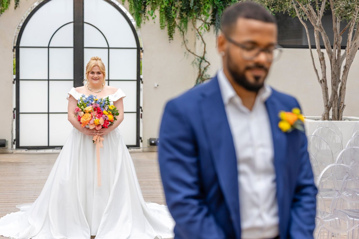 Bride in white wedding dress holding colorful bouquet while groom in blue suit stands in foreground at outdoor venue.