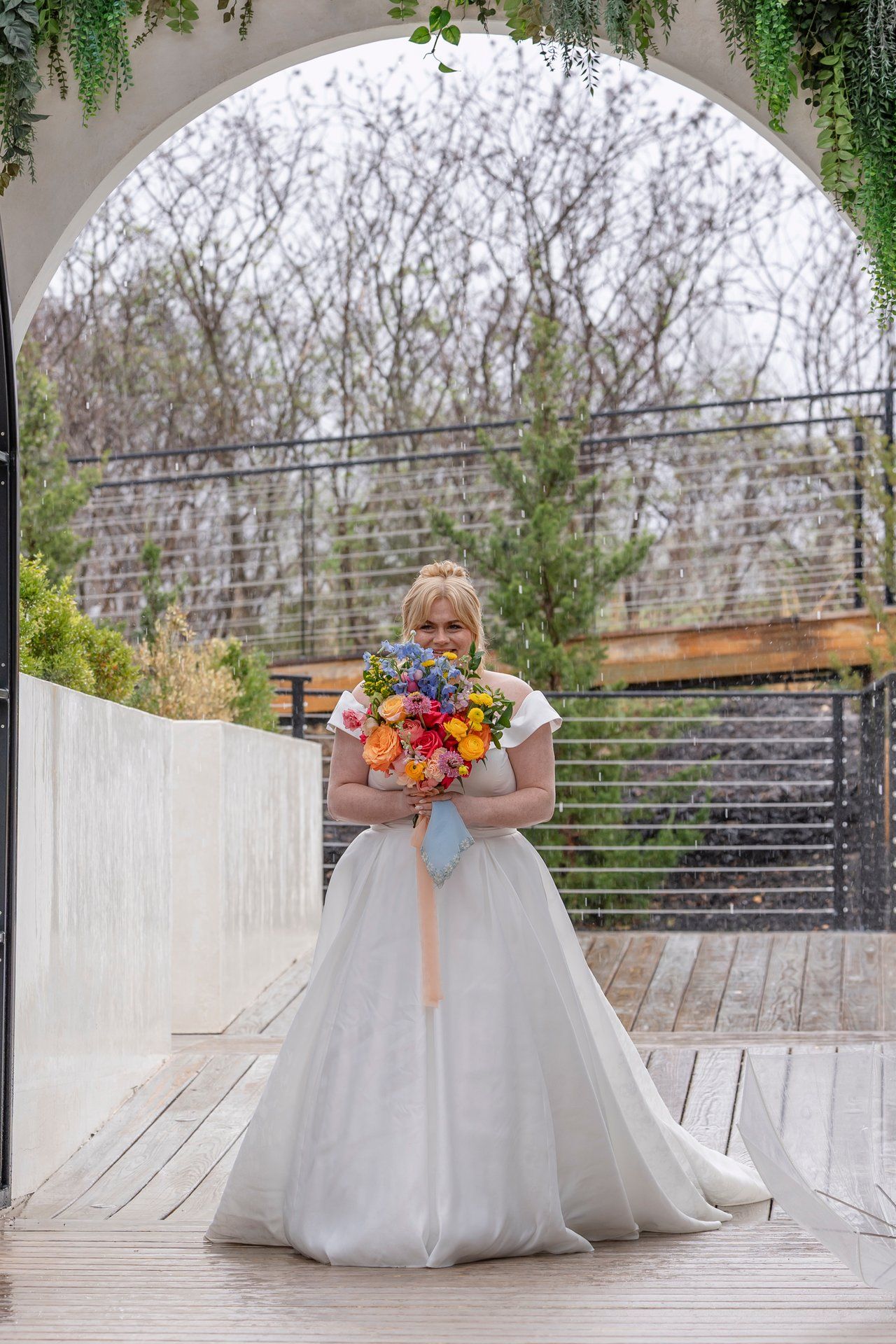 A bride in a flowing white wedding gown holds a vibrant fall-colored bouquet under an arched garden walkway.