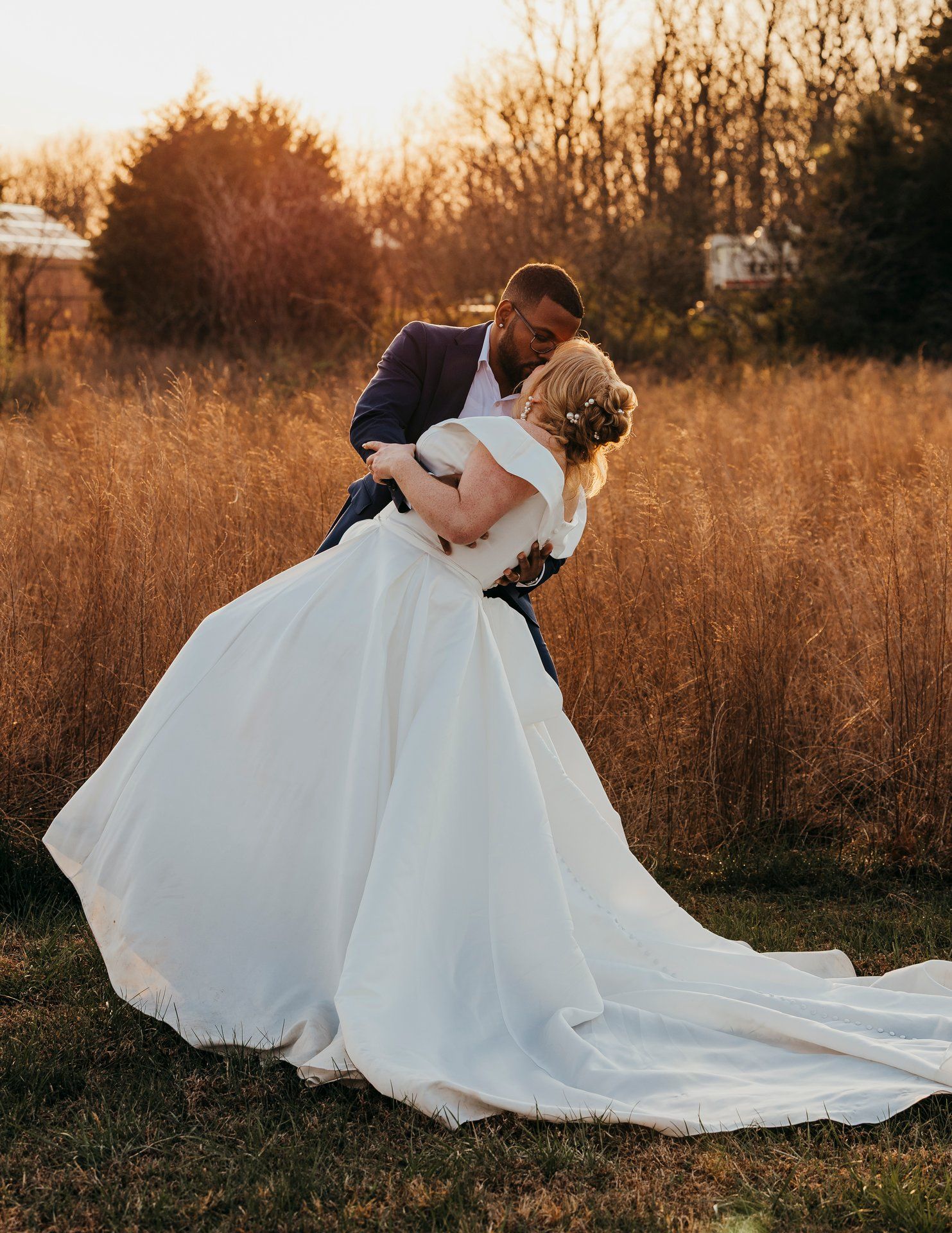 Wedding couple shares intimate moment in golden field at sunset with bride in flowing white gown.