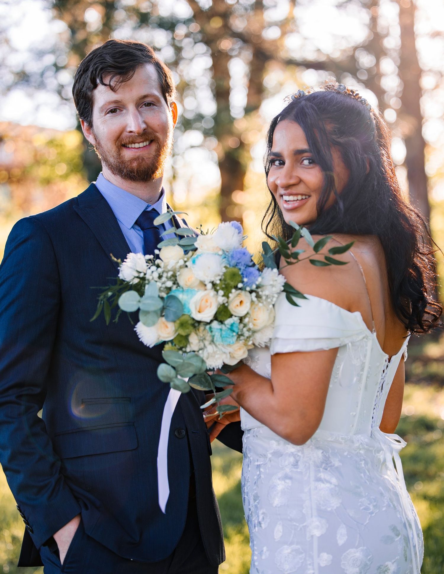 Joyful newlyweds pose together outdoors holding a beautiful white and blue floral bouquet.
