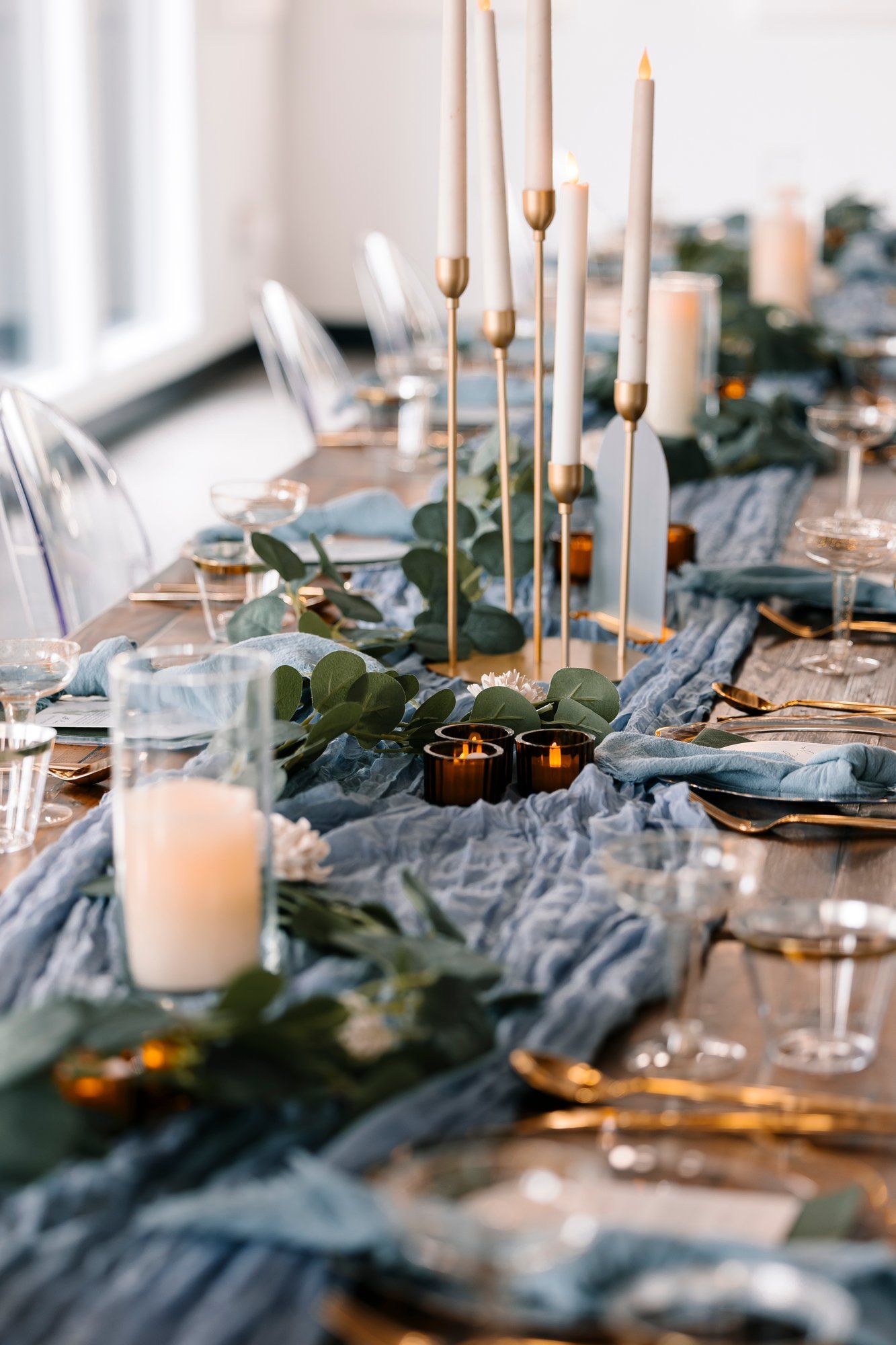 Elegant wedding table setting with dusty blue fabric, white candles, and eucalyptus greenery in a soft focus style.