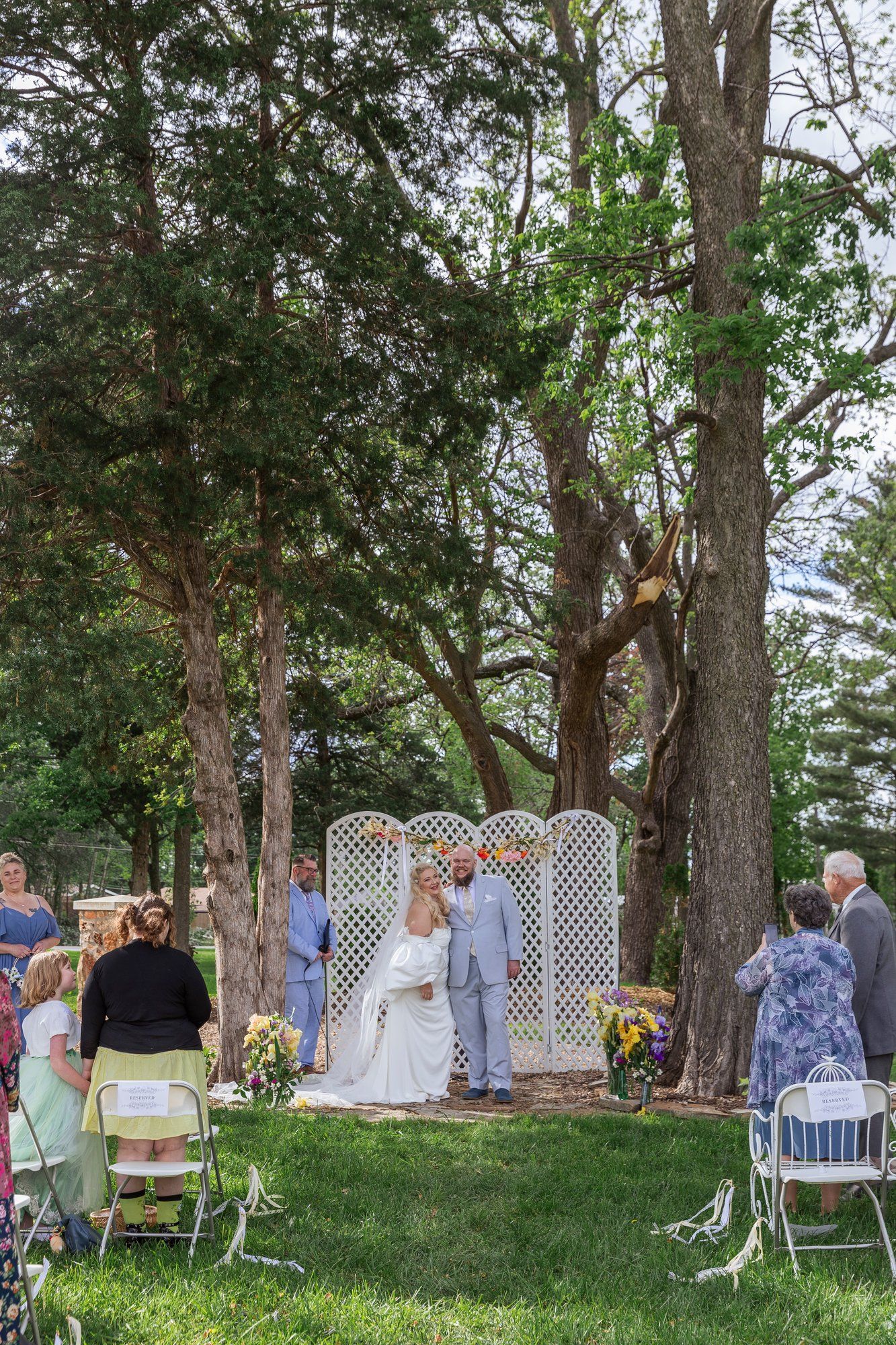 A series of outdoor wedding ceremony shots with guests seated under tall trees and white lattice decorations.