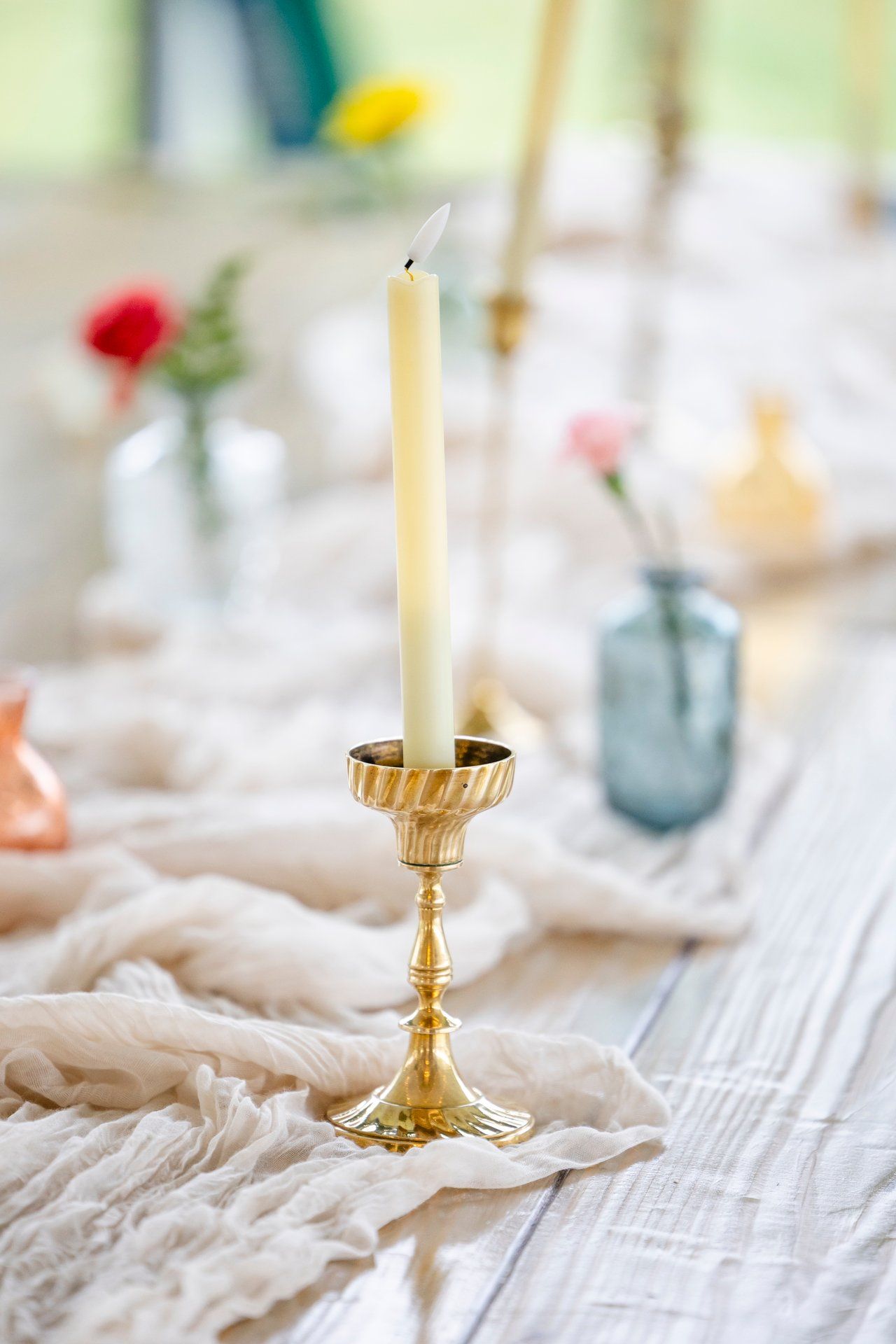 A lit white taper candle in an elegant brass candlestick holder with blurred glass vases in the background.