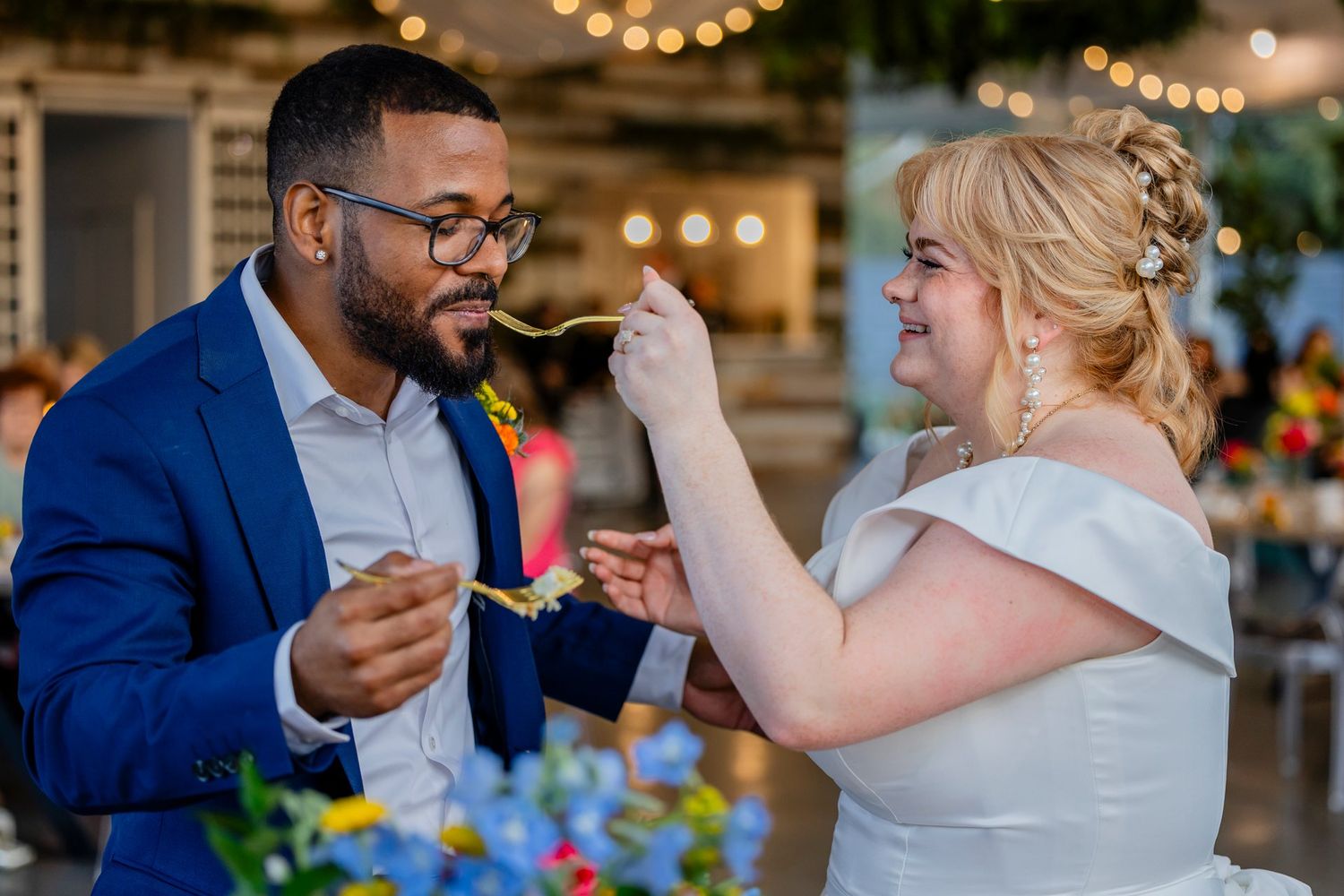 A couple shares cake at their wedding reception surrounded by twinkle lights and floral decor.