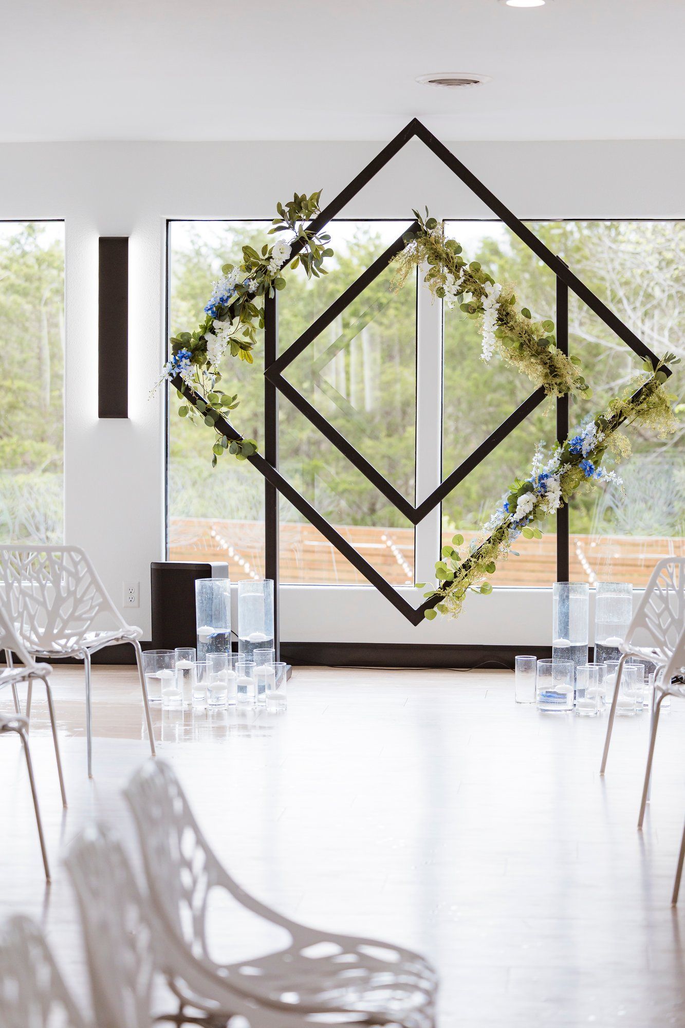 Modern geometric wedding arch decorated with florals stands before large windows in a minimalist white ceremony space.