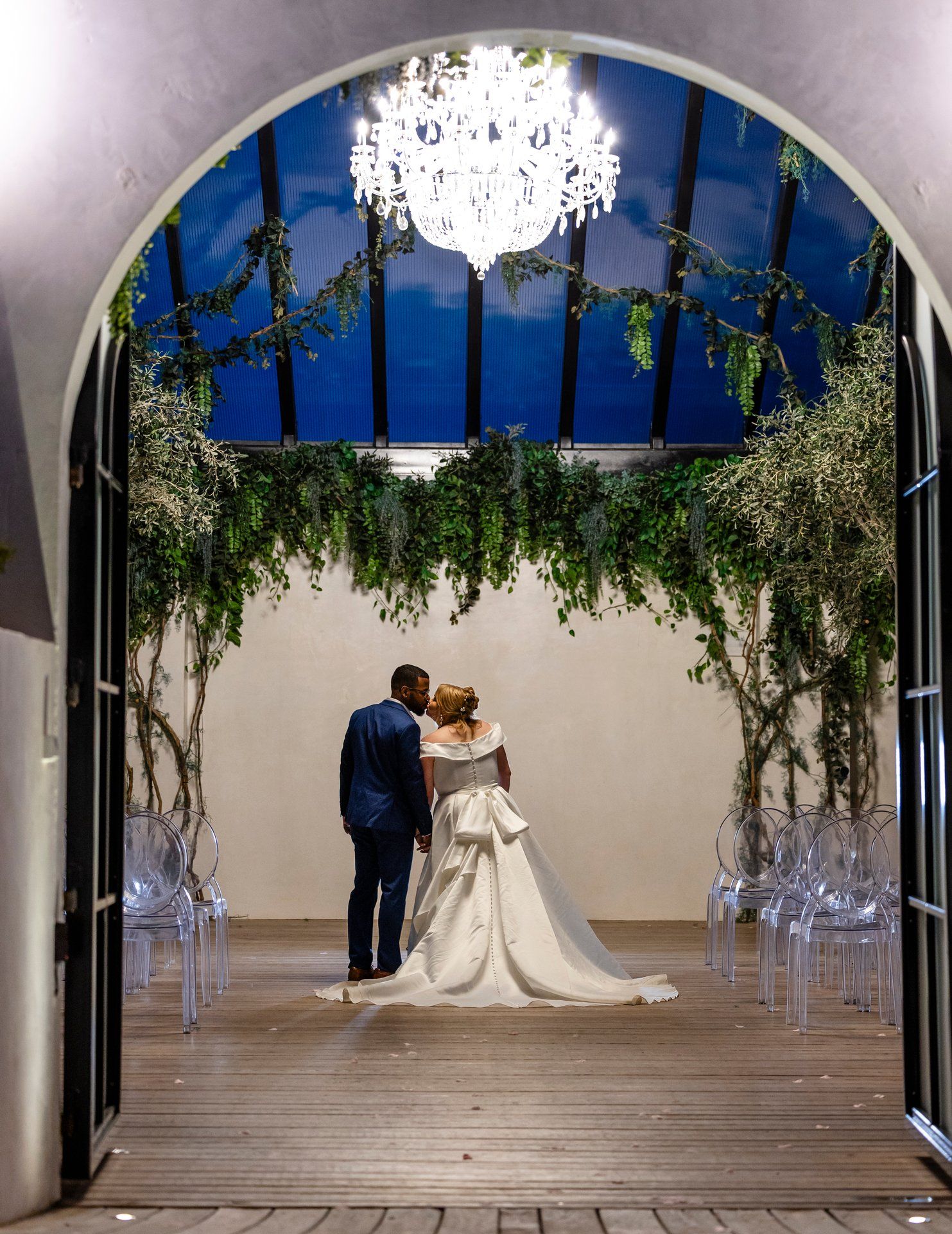 A wedding couple captured under an arched doorway with hanging greenery and a crystal chandelier.