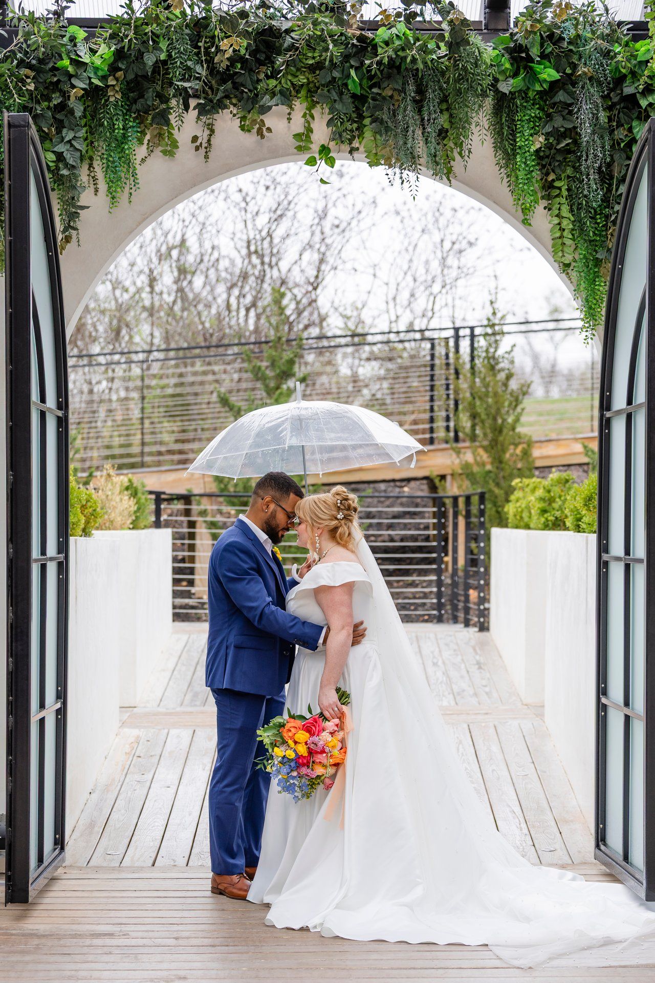 Bride and groom share a moment under a greenery-adorned archway with clear umbrella.