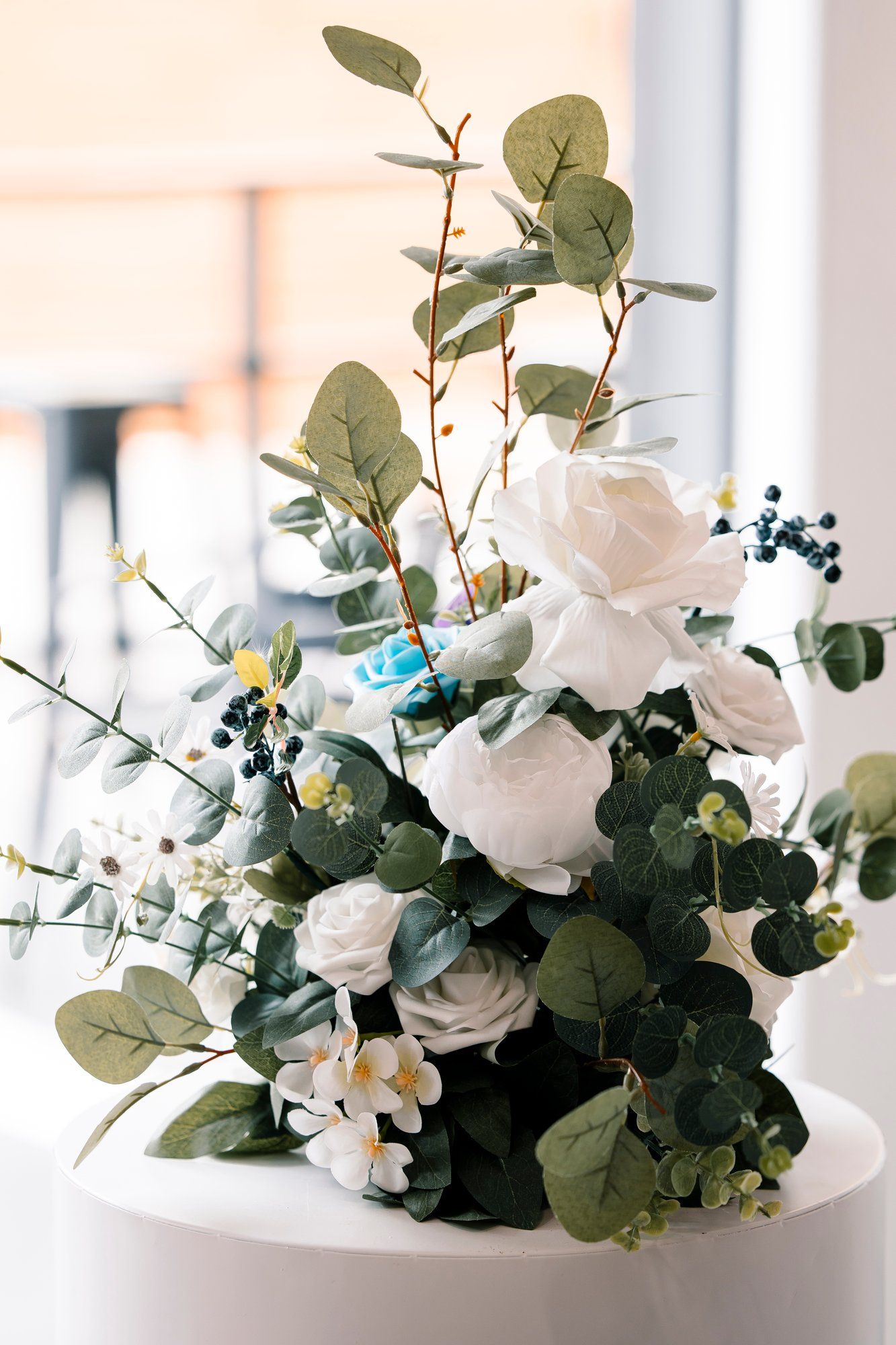 Elegant floral arrangement featuring white roses, eucalyptus leaves and small blue berries in a white vase with soft lighting.