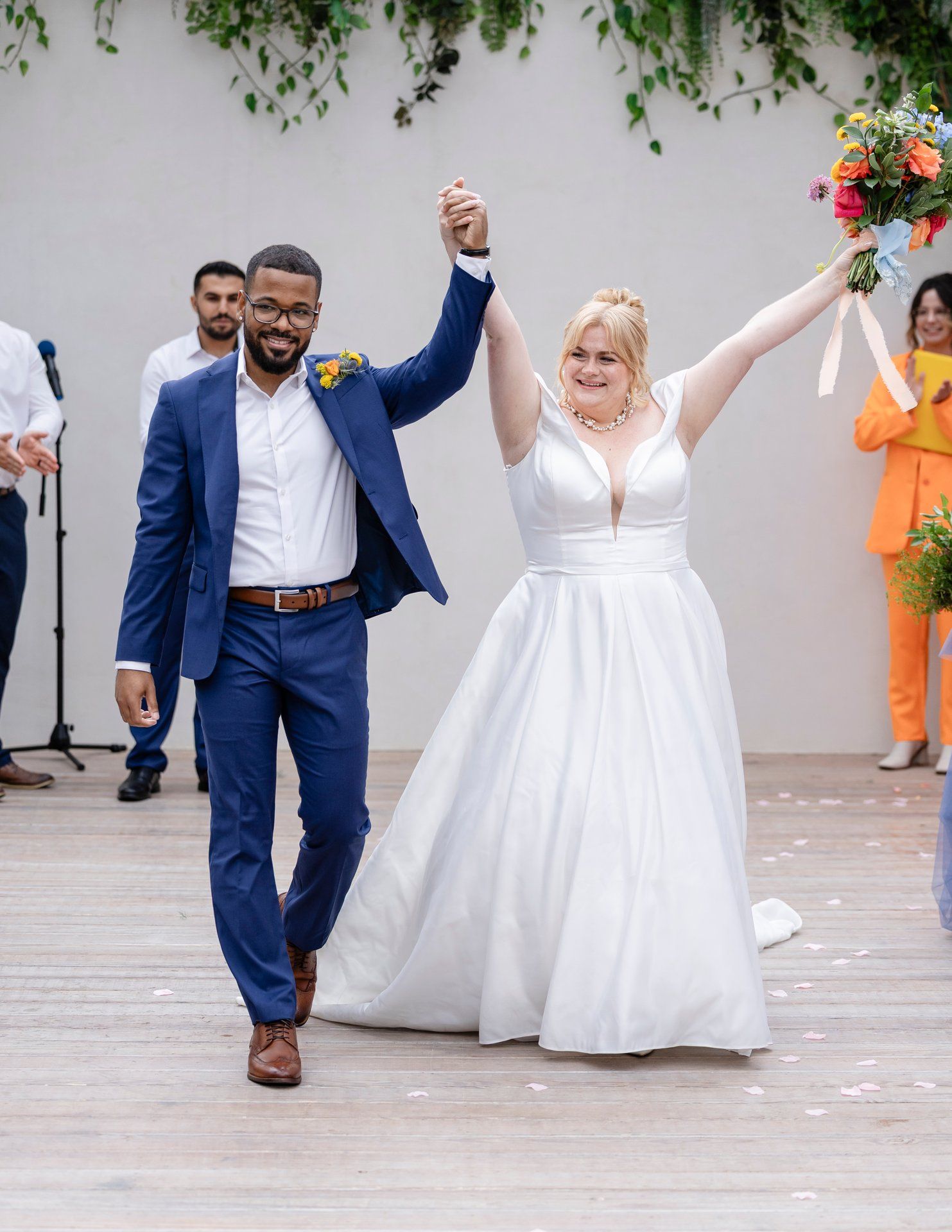 A couple celebrates at their wedding ceremony, holding hands raised in joy while wearing blue suit and white gown.