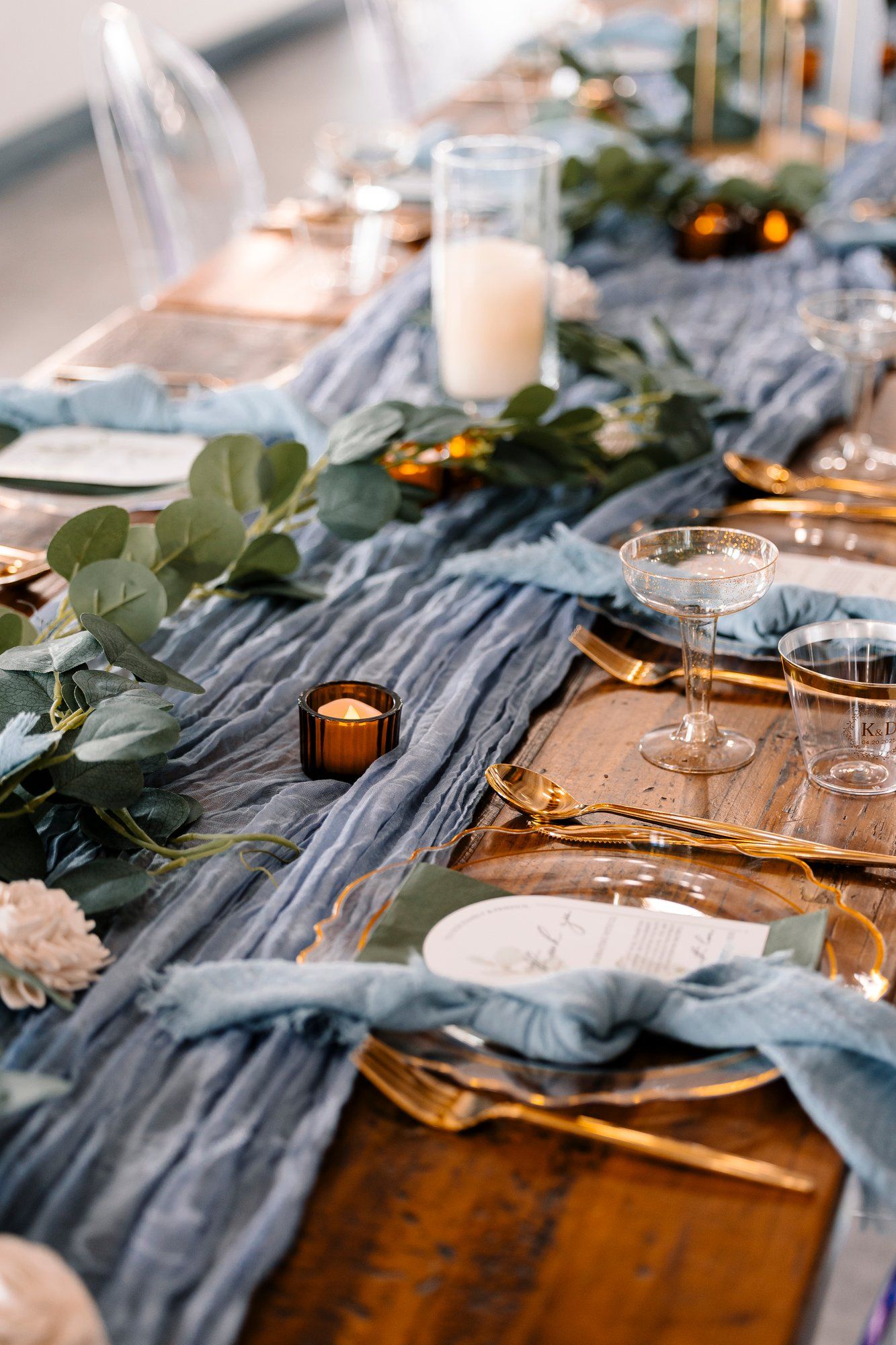 An elegant table setting with blue gauzy runner, eucalyptus leaves, gold cutlery and glowing candles for a romantic dinner.