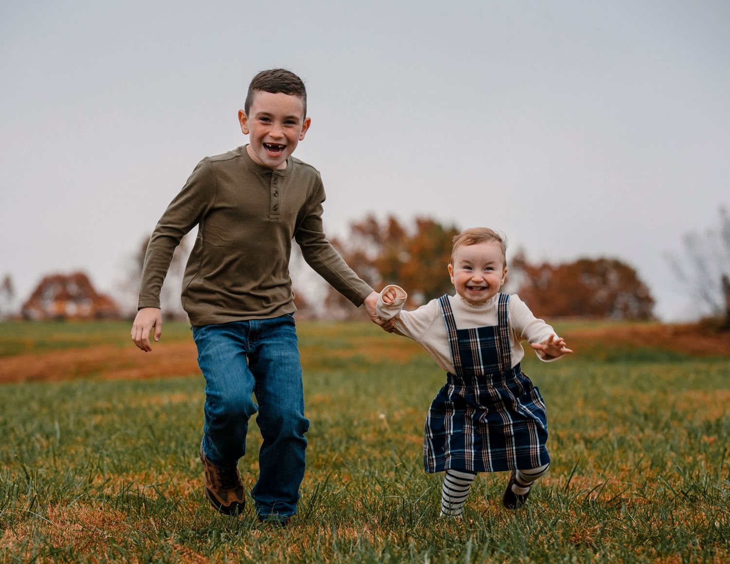 In Missouri, Two children run and laugh together across a grassy field on an autumn day with trees and a house in the background.