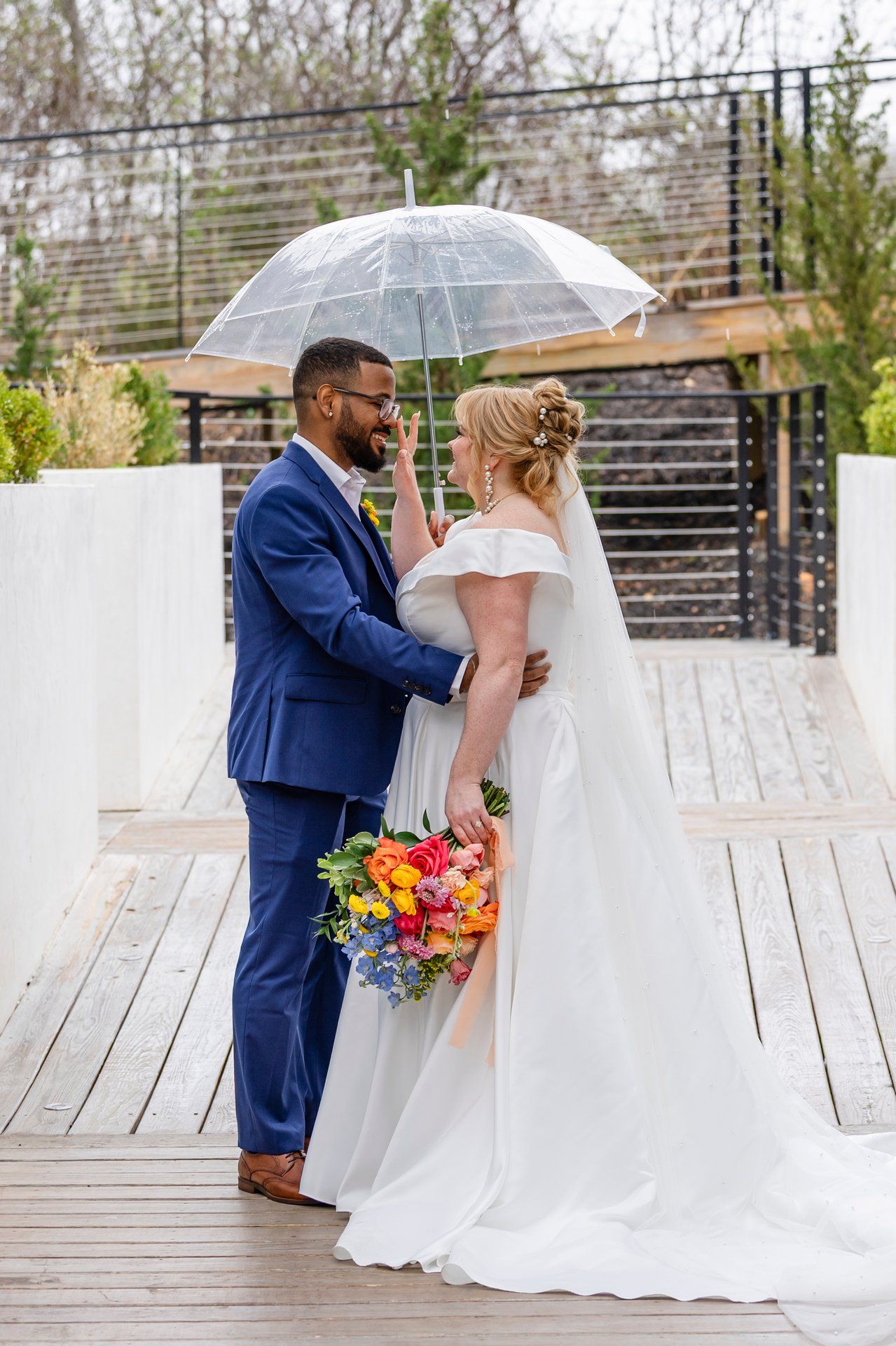 Newlyweds share an intimate moment under a clear umbrella on a wooden deck.