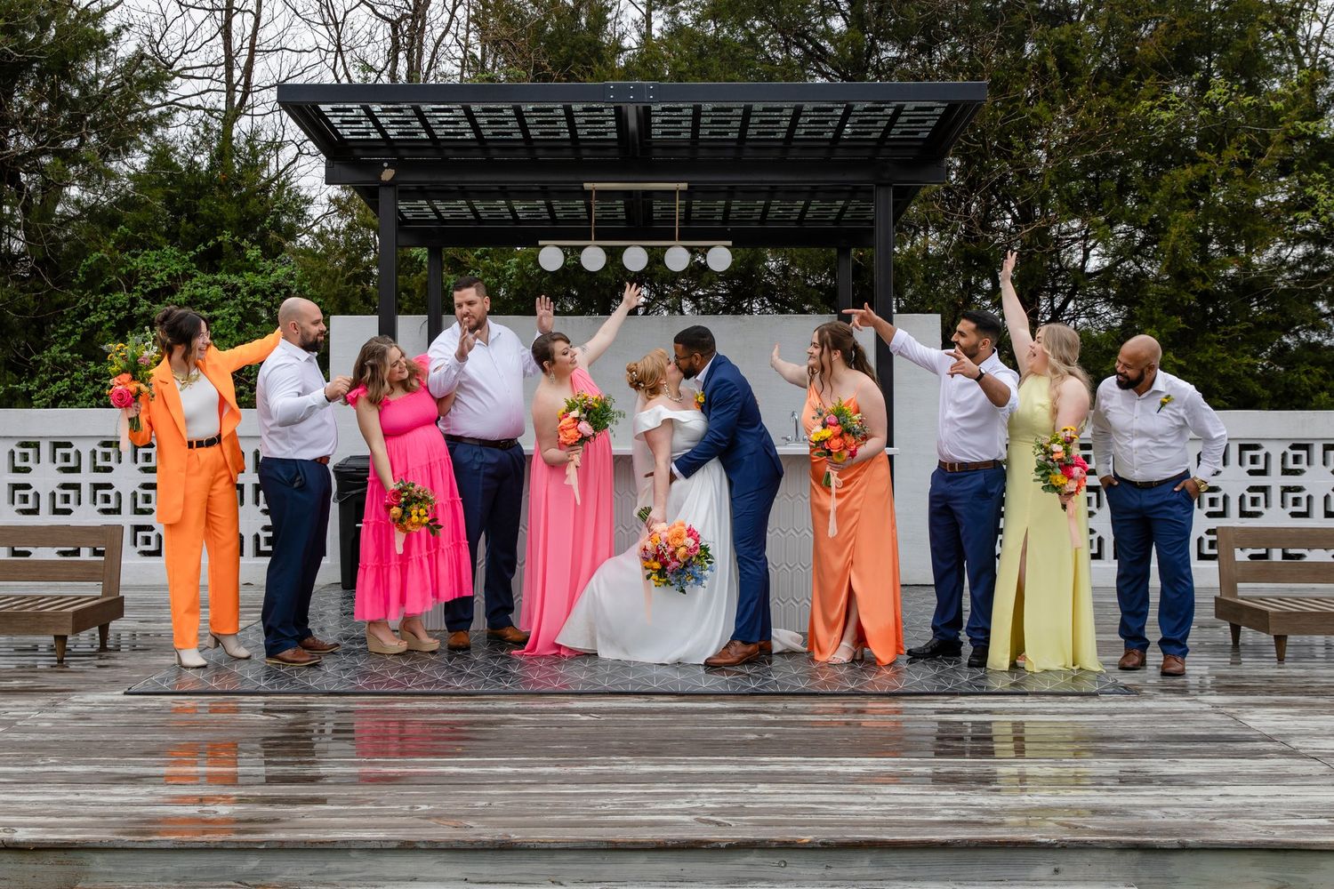 A wedding party in colorful attire poses on outdoor steps under a covered pavilion.