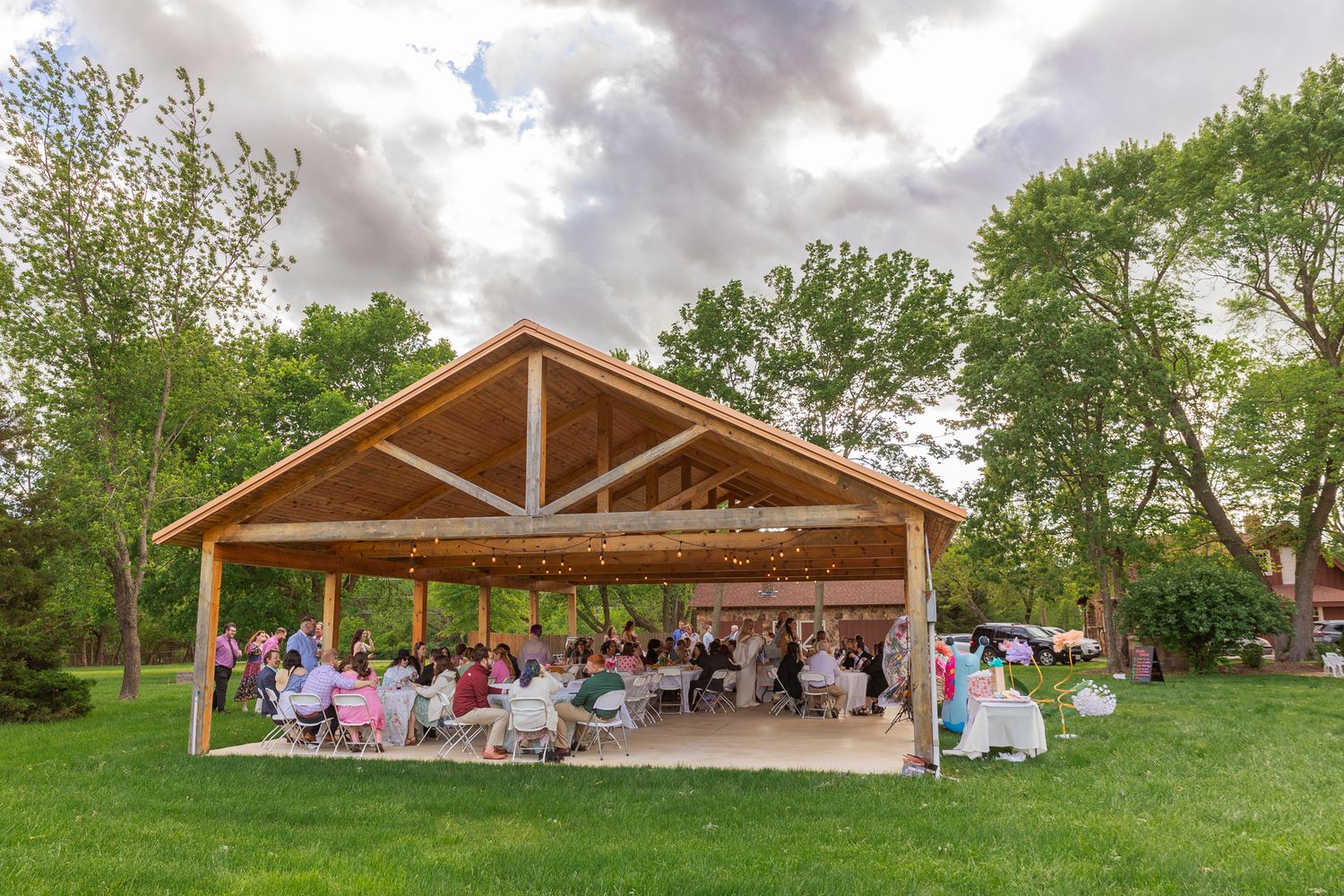 A rustic wooden pavilion filled with wedding guests seated at tables on a lush green lawn under cloudy skies.