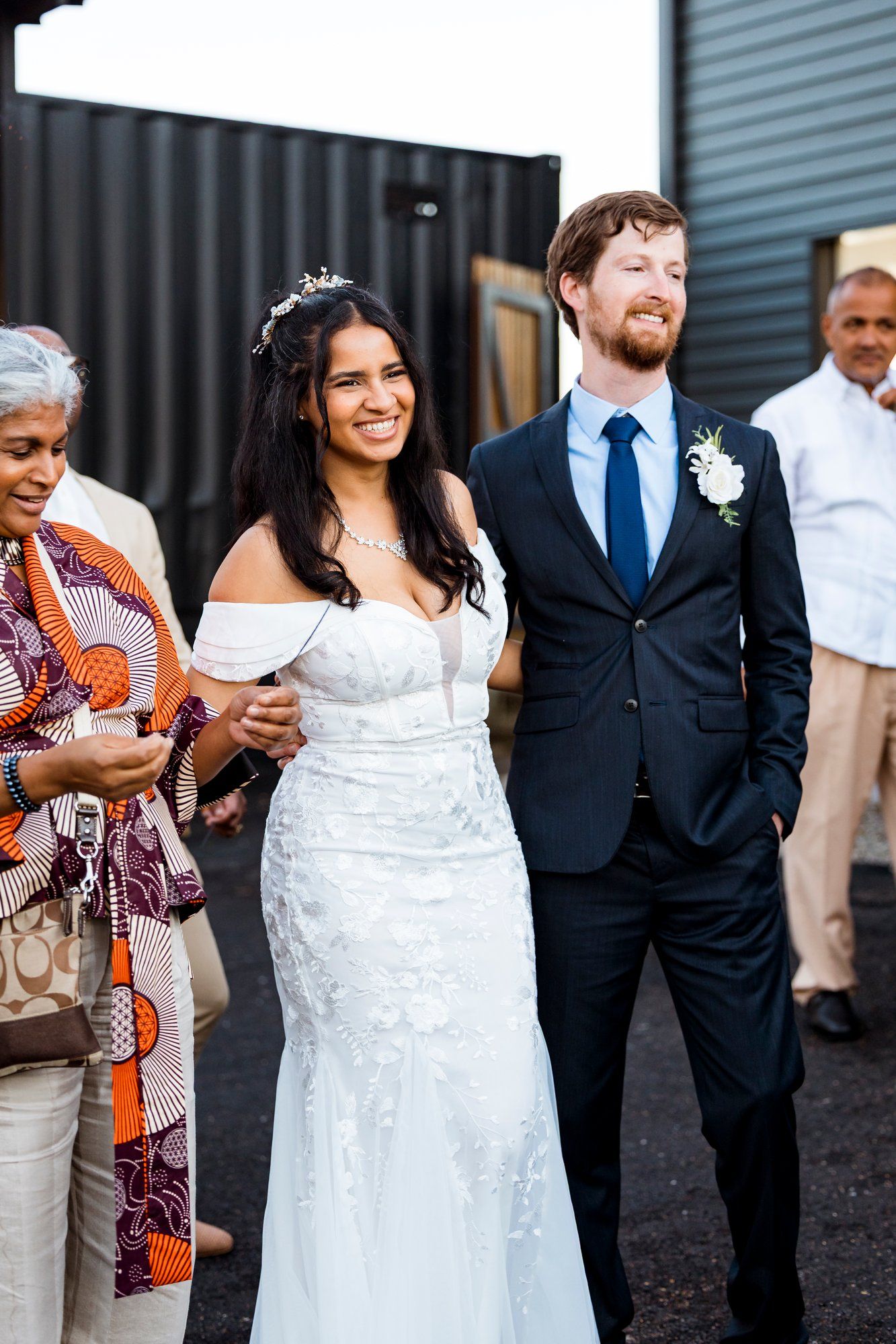 Wedding party stands together outdoors, with couple in white dress and navy suit next to guest in colorful patterned dress.