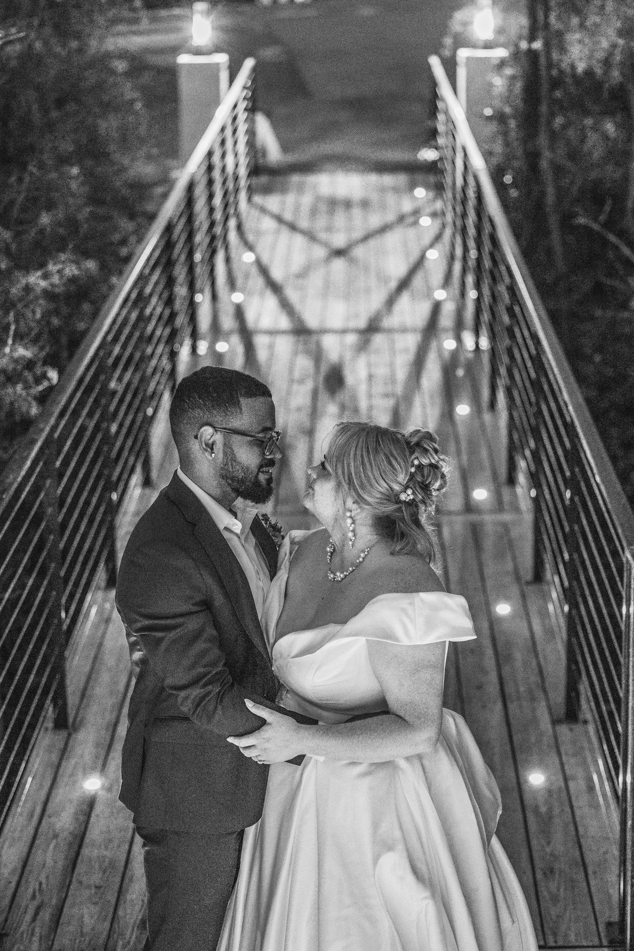 Romantic black and white wedding portrait of couple standing on illuminated suspension bridge at night.