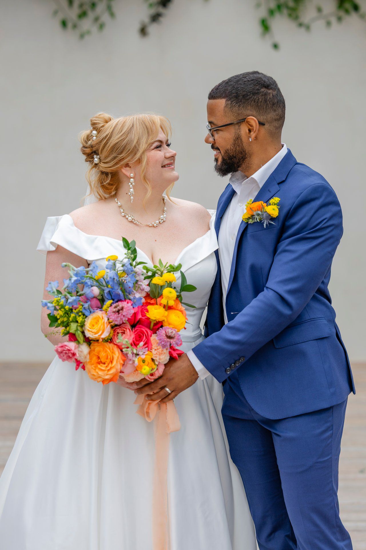 Bride holds vibrant multicolored bouquet while sharing a tender moment with groom in navy suit.