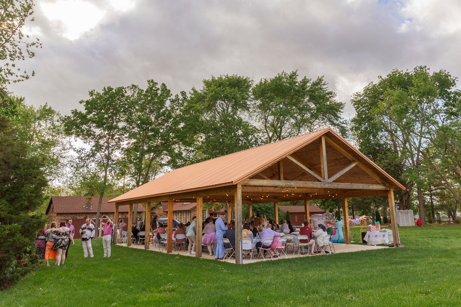 Large open-air pavilion with exposed wooden beams hosts outdoor gathering on lush green lawn under cloudy skies.