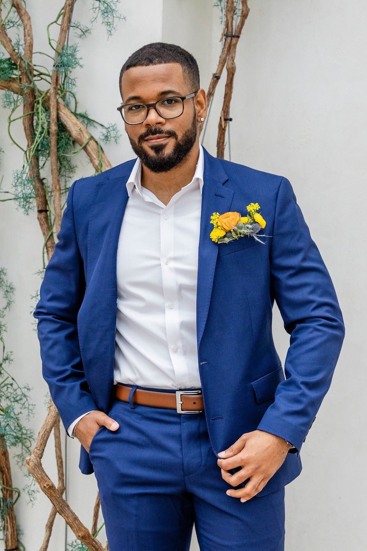 A groom in a tailored blue suit and white shirt poses confidently against a white wall with climbing vines.