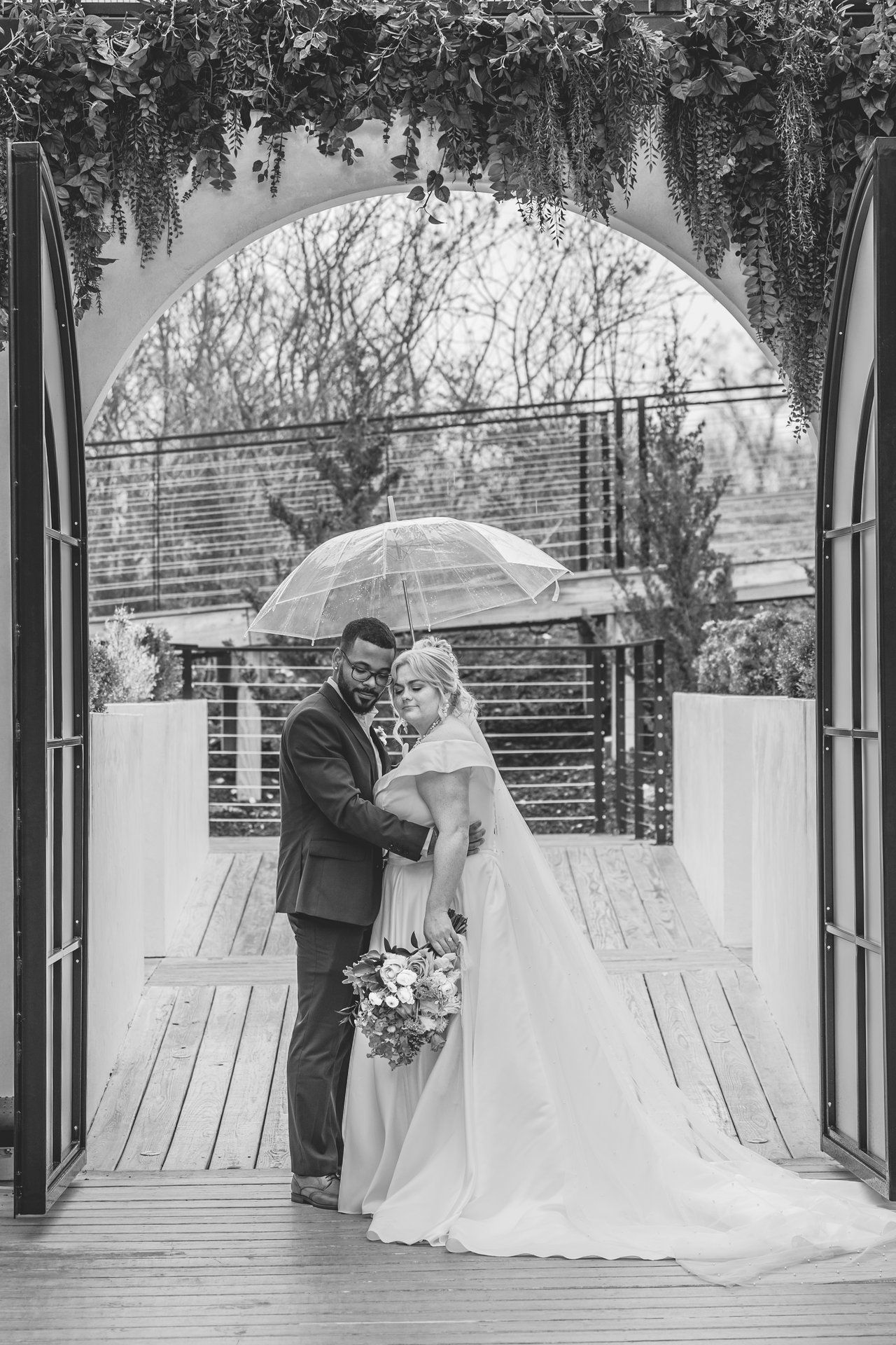 A black and white photo of newlyweds sharing an intimate moment under an umbrella in a garden archway.