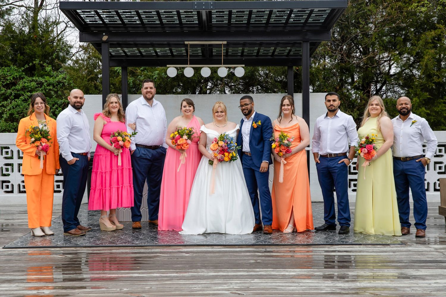 Wedding party lined up in vibrant orange and pink dresses on rainy day.
