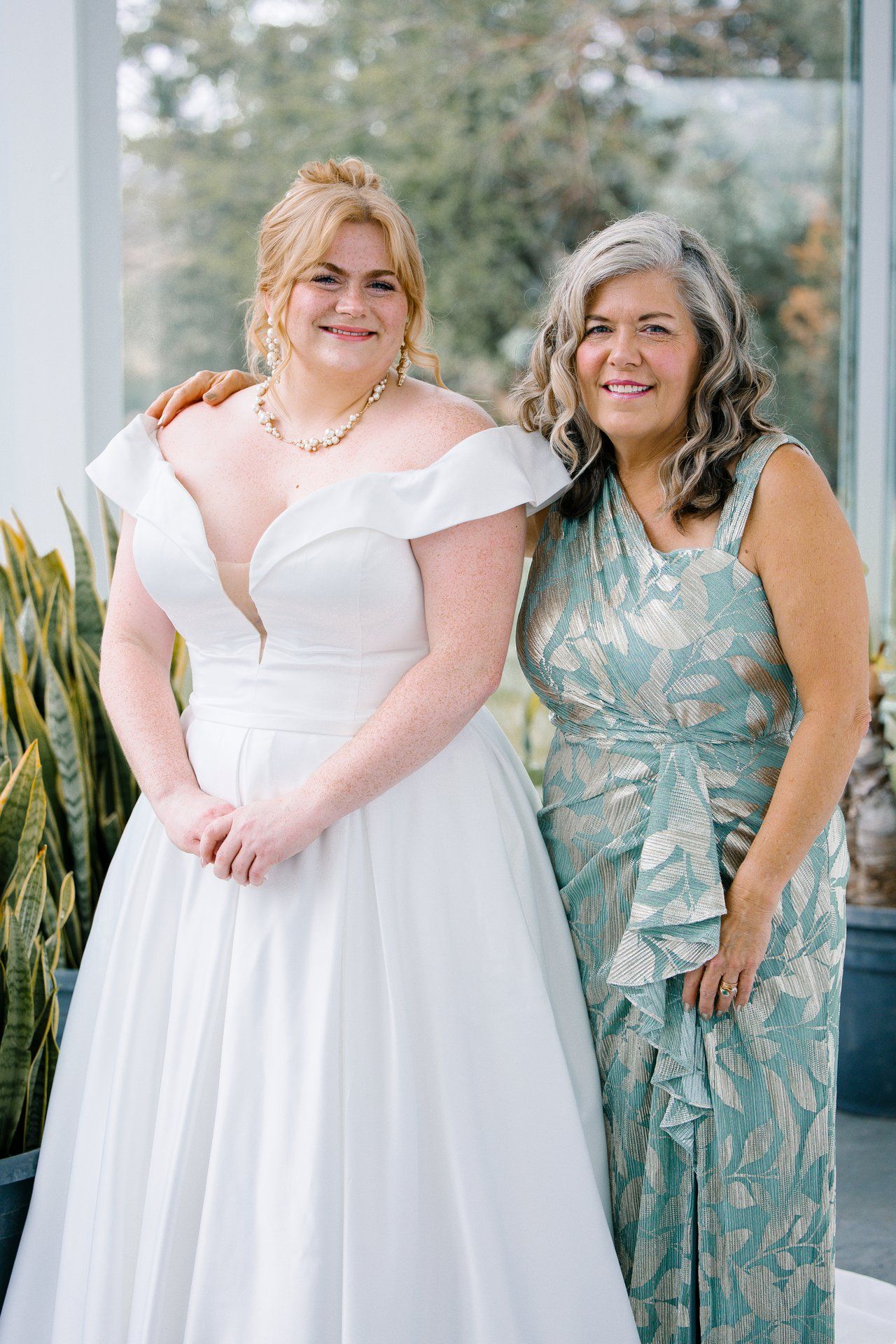 A joyful wedding photo showing a bride in a white gown and guest in a blue-green floral dress.