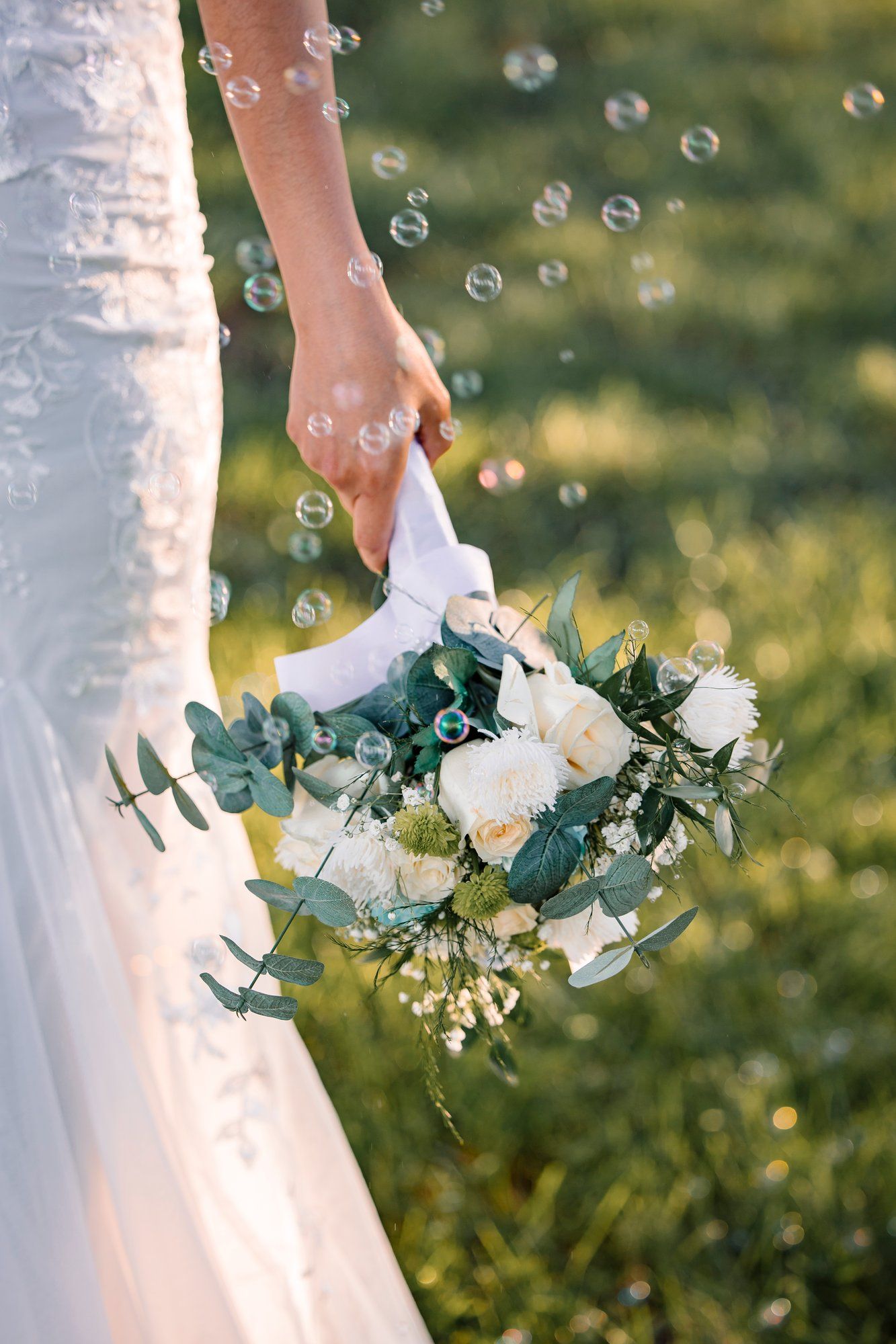White wedding dress and floral bouquet details with delicate soap bubbles floating in sunlit garden setting.