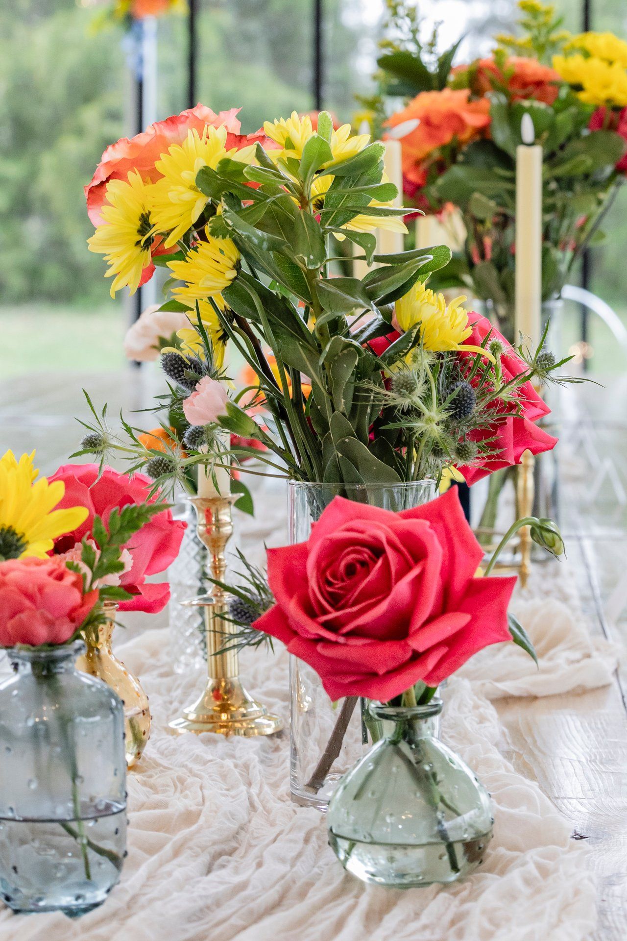 Beautiful centerpiece arrangement with pink roses, yellow sunflowers and mixed flowers in small glass vases on textured fabric.