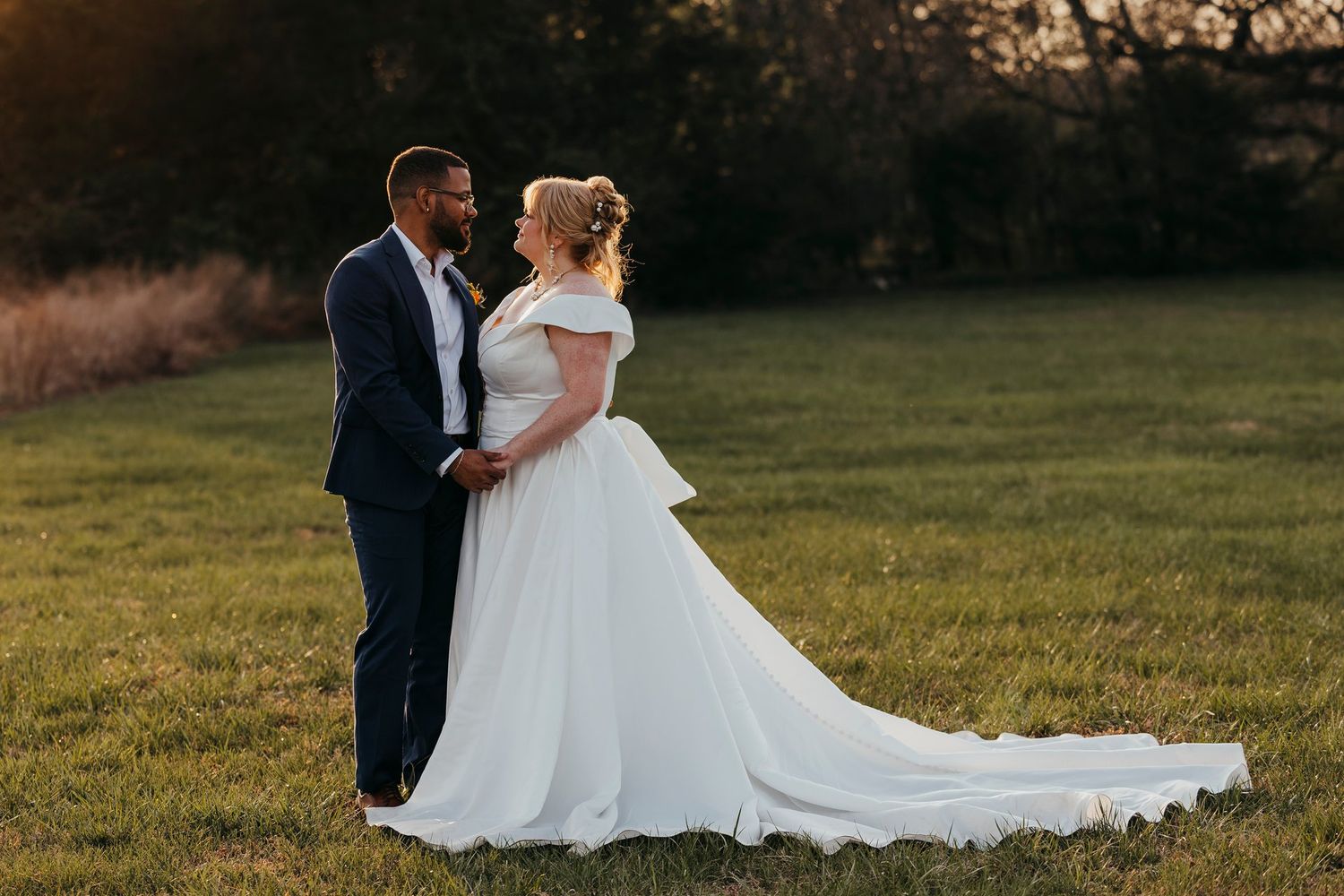 A newlywed couple stands together in a field at sunset.