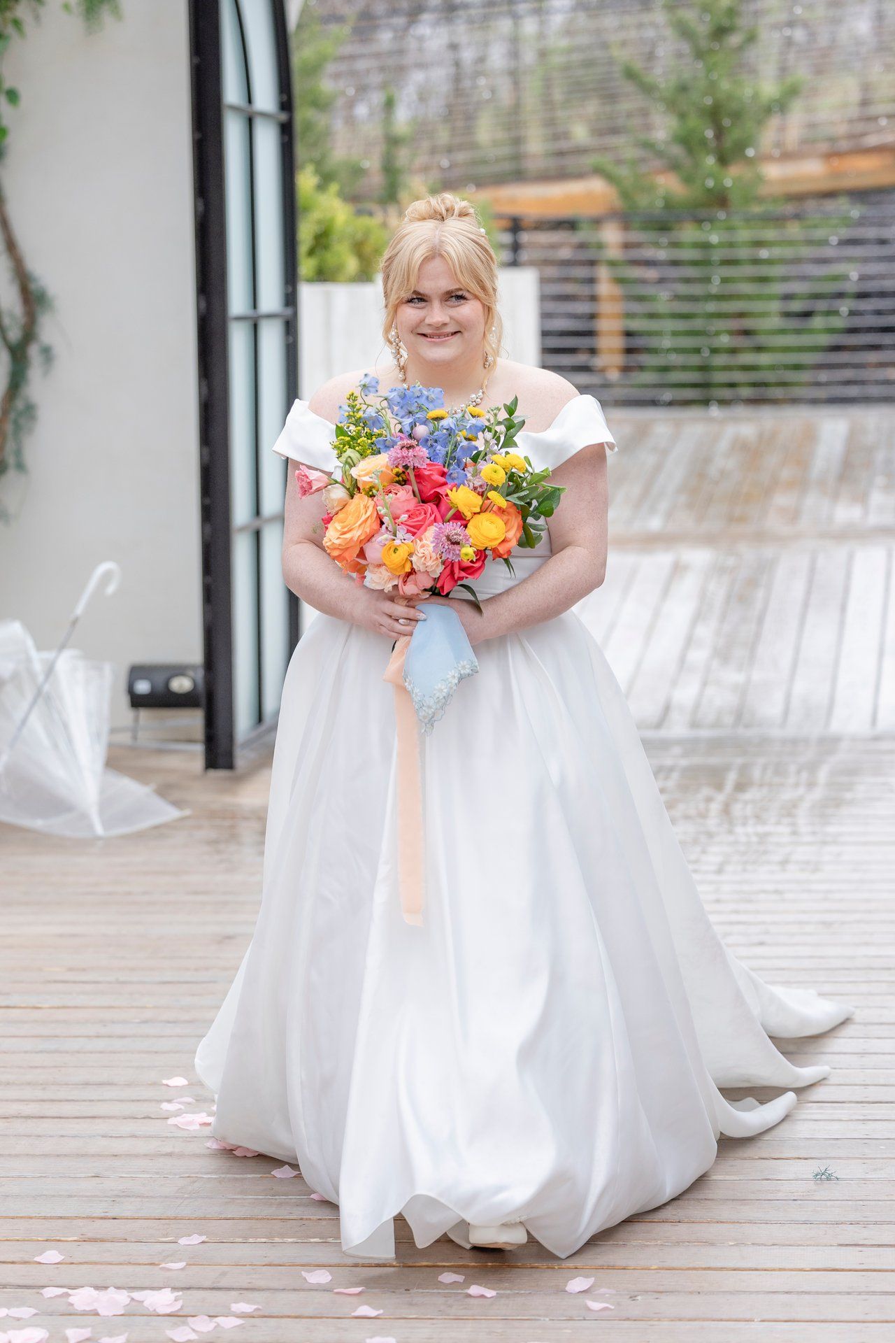 A bride in a flowing white wedding dress holds a vibrant bouquet of colorful flowers on a wooden deck.