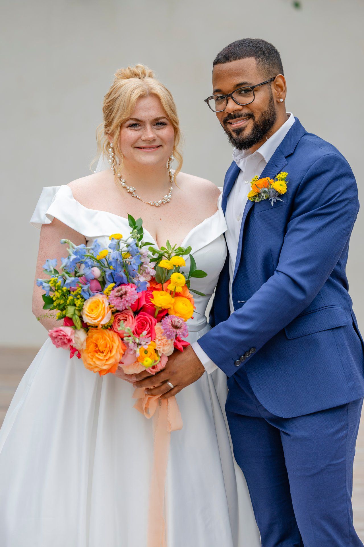 Newlyweds pose with bright floral bouquet against white wall with climbing vines.