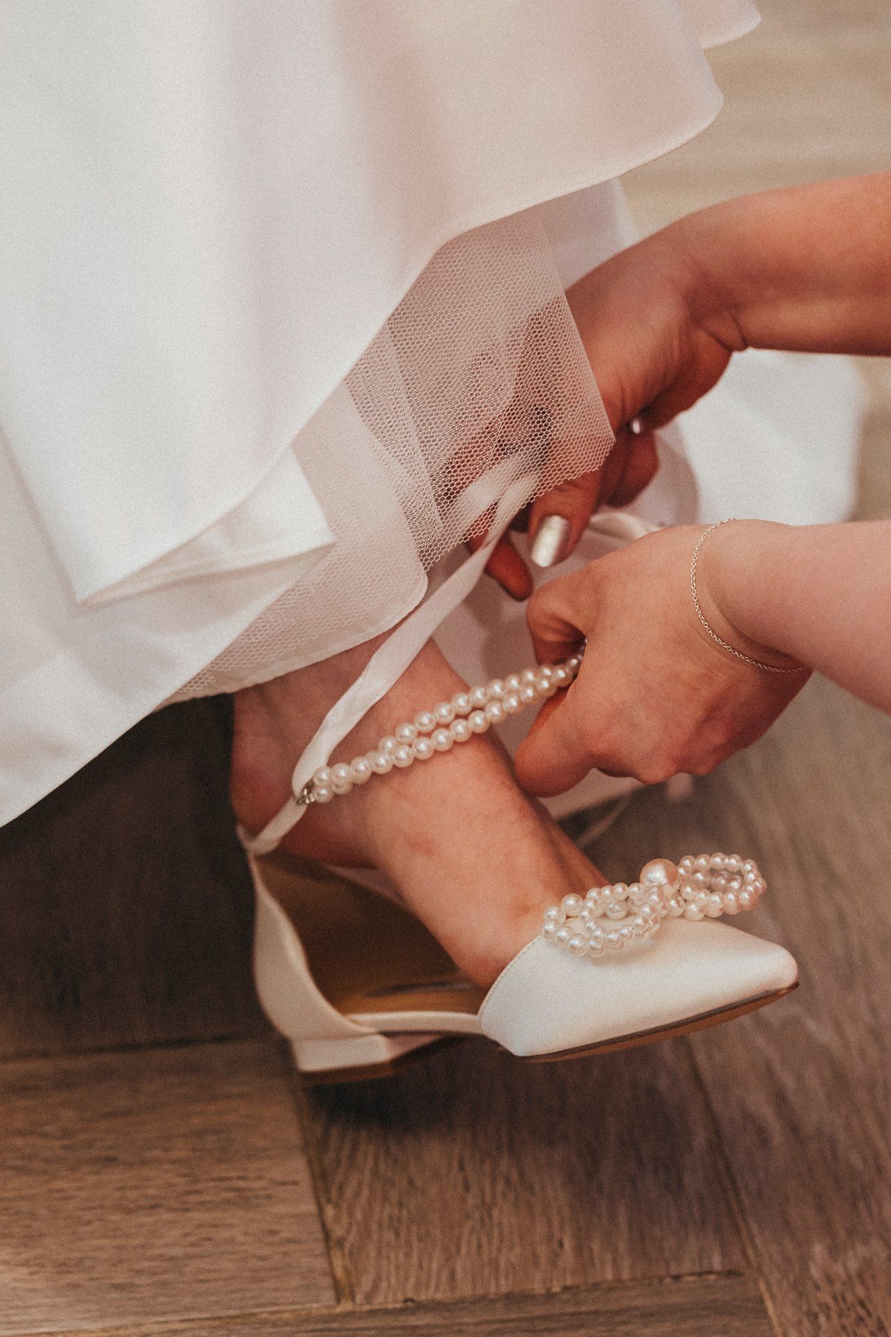 Hands fastening delicate pearl straps on a white bridal shoe on hardwood floor.