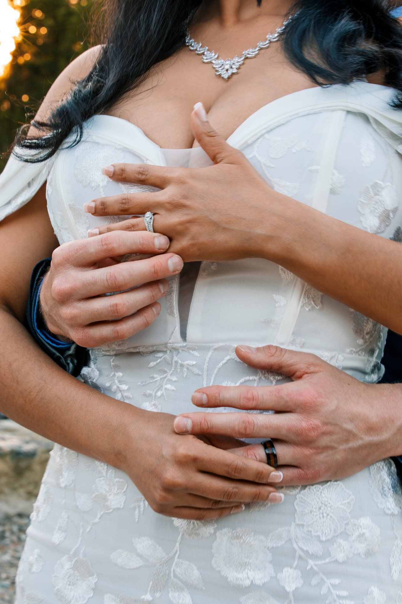 Close-up detail of hands with wedding rings against a white lace wedding dress and sparkly necklace.