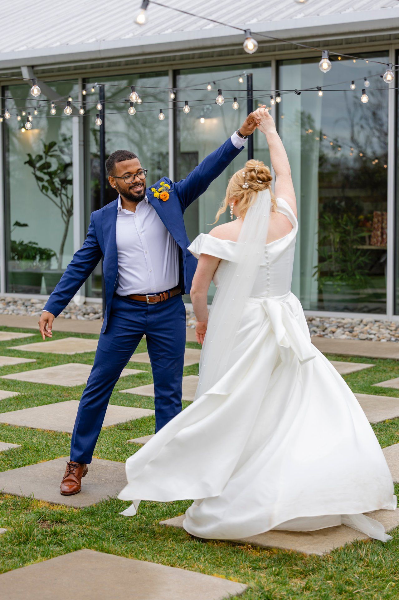 Happy newlyweds dancing joyfully outdoors with string lights and glass windows in the background.