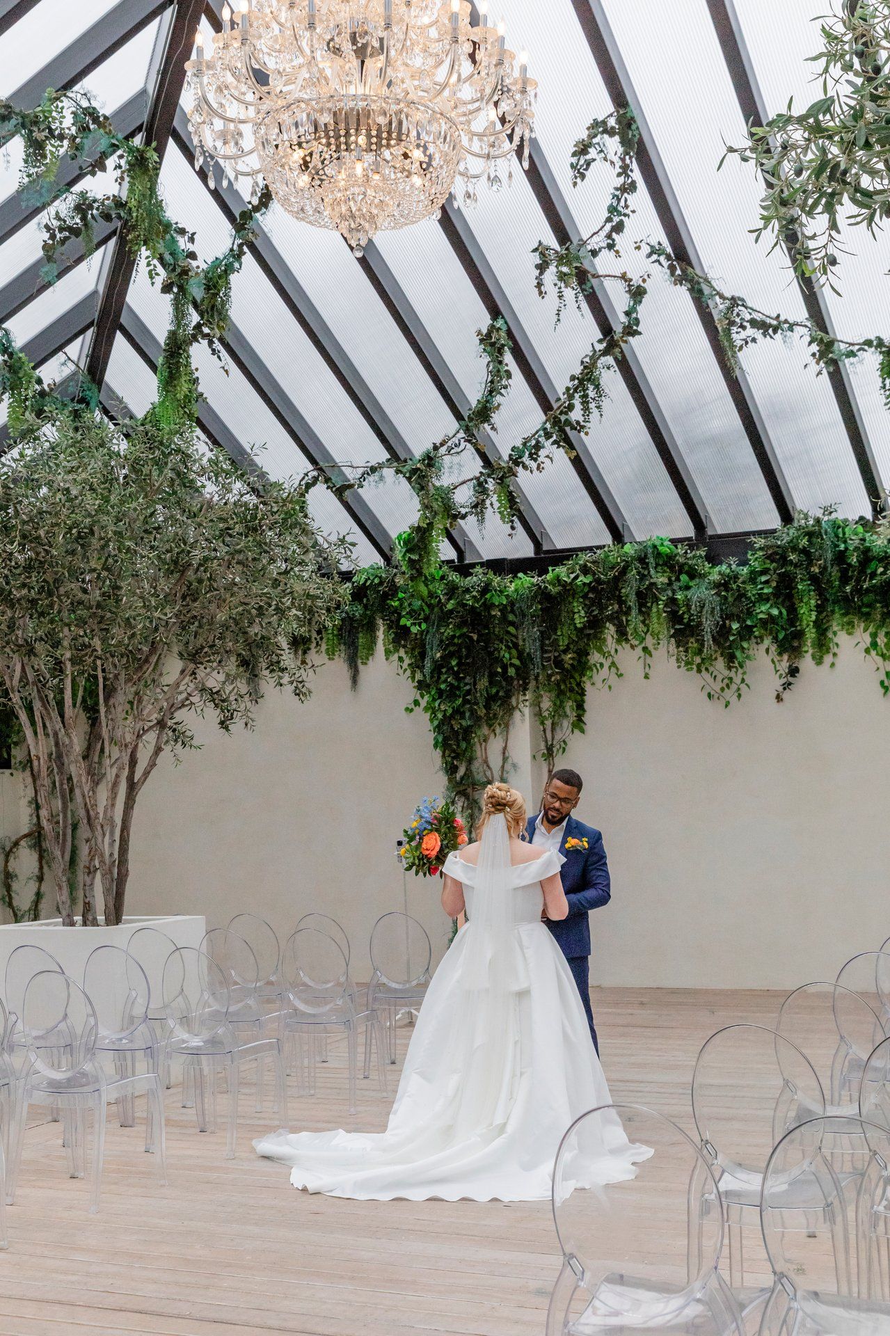A wedding ceremony setup in a greenhouse with chandelier, clear chairs and greenery draped from glass ceiling.