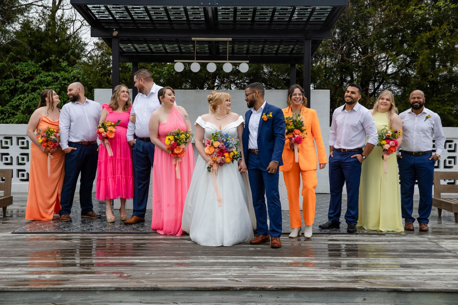 Wedding party in colorful attire poses together on rainy day under pergola.