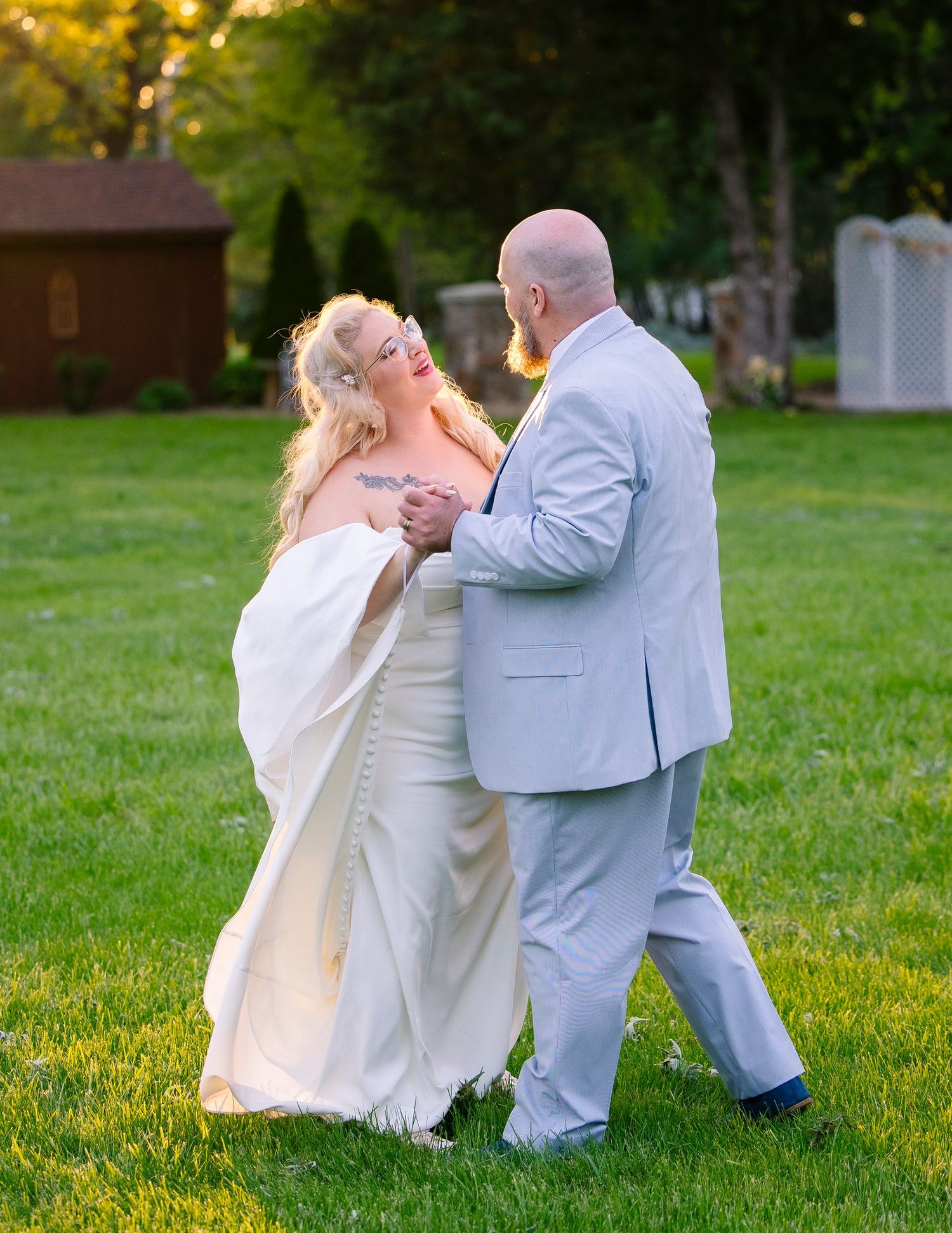 A couple in light colored formal wear dance together on a grassy lawn during a romantic outdoor celebration.