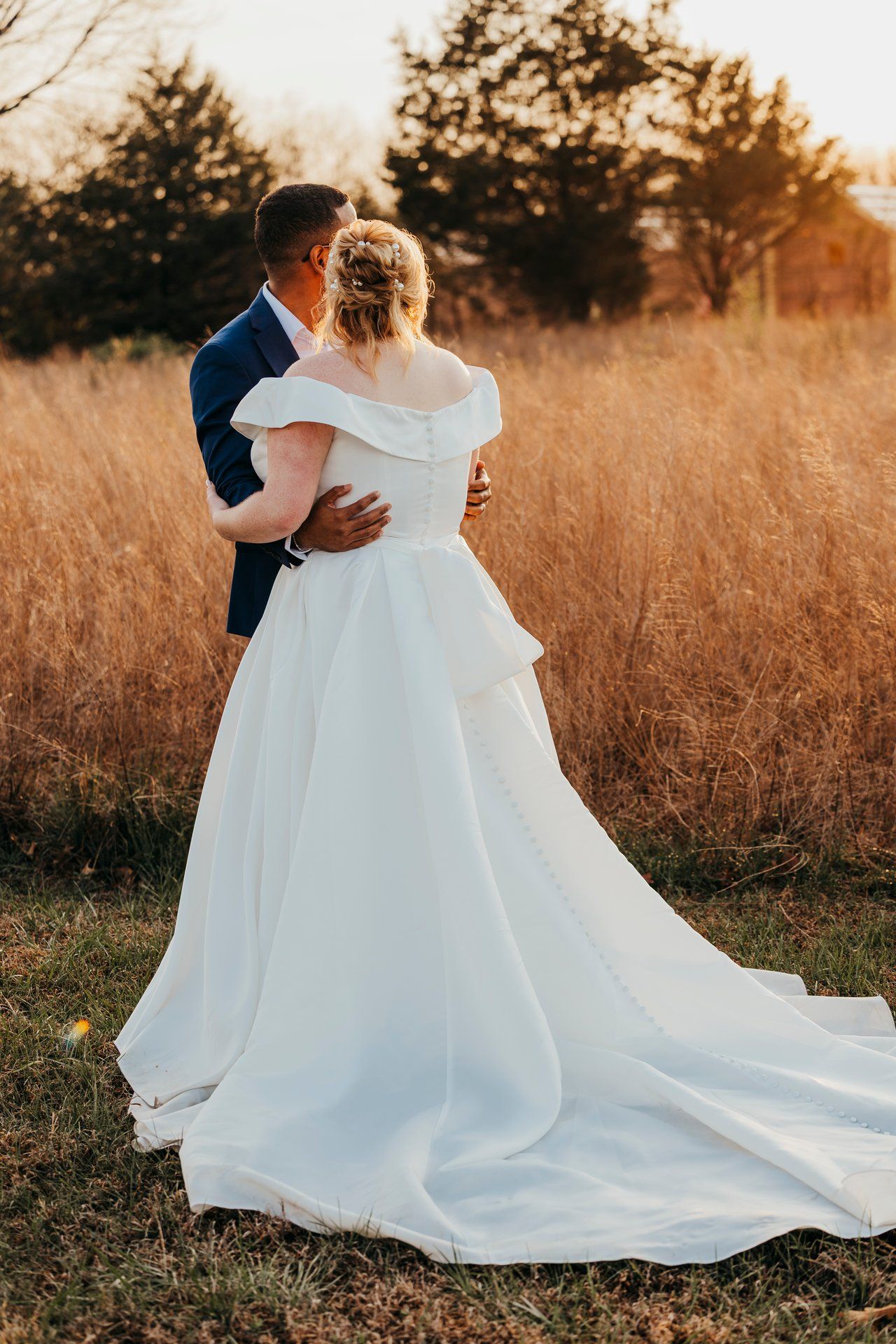 Bride and groom embracing in golden sunset field with flowing wedding dress.