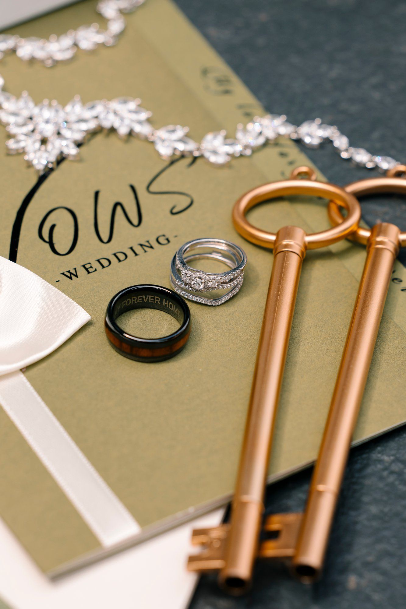 Wedding rings and copper vintage keys arranged on an olive green invitation with delicate white lace trim details.
