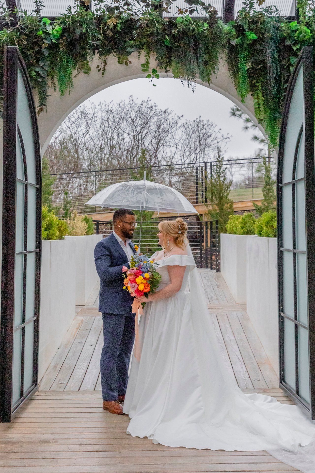 Bride and groom stand under floral archway holding clear umbrella on rainy wedding day.