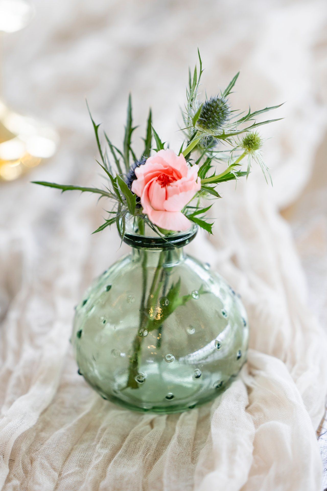 A pink rose and thistle arrangement in a textured green glass vase sits on white fabric.