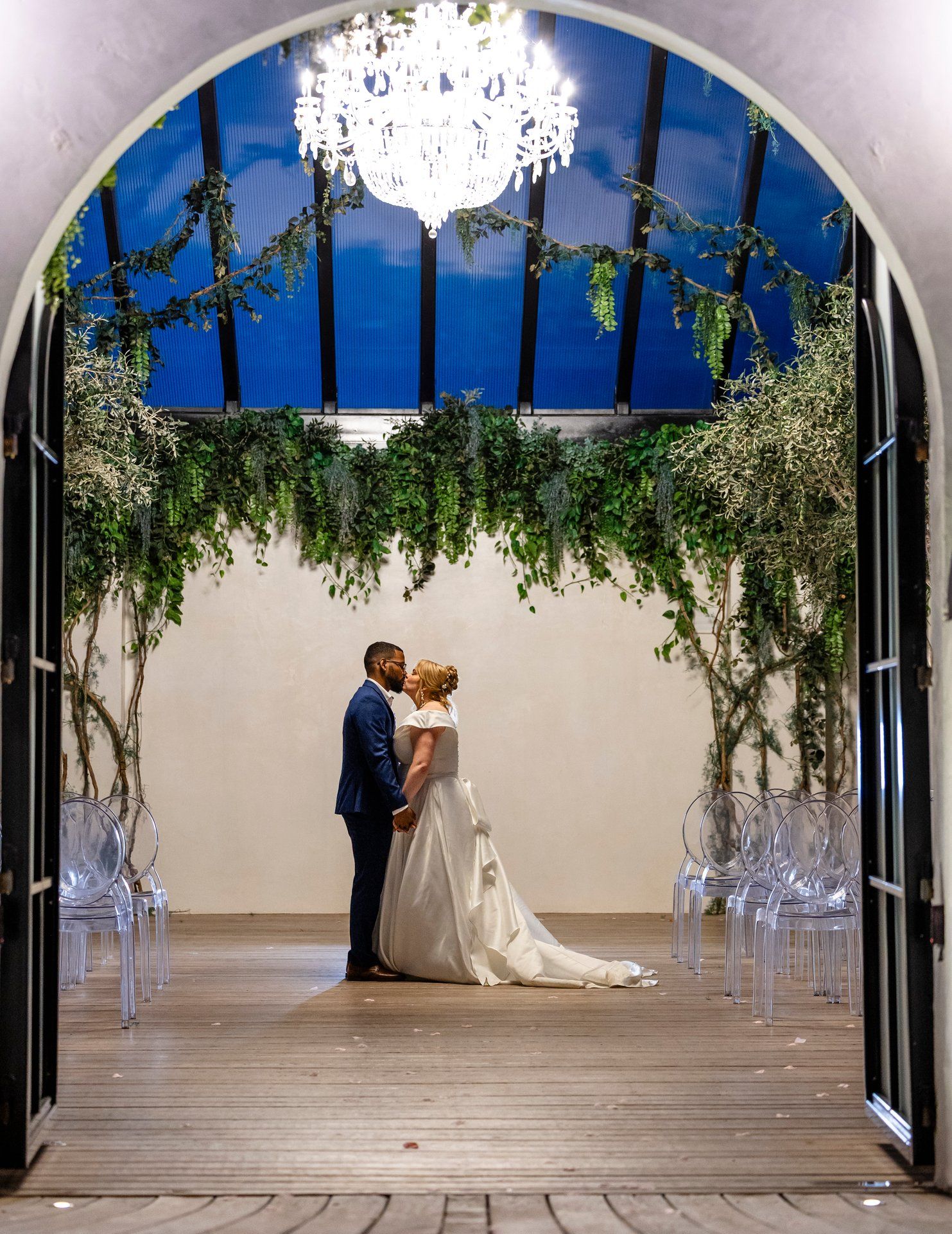 Elegant wedding couple kissing under crystal chandelier and floral arch.
