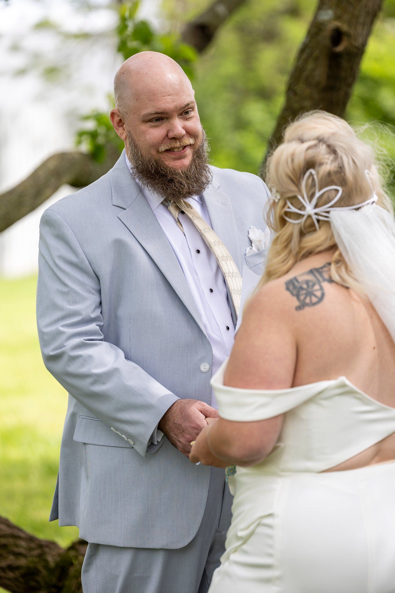 A groom in a light gray suit and beard smiles lovingly at his bride during an outdoor wedding ceremony in a garden setting.