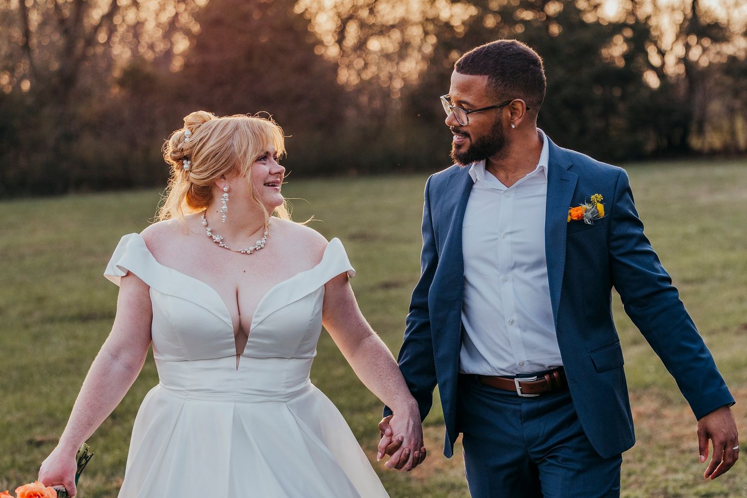 Newlyweds walk hand in hand through grassy field during sunset in elegant wedding attire.